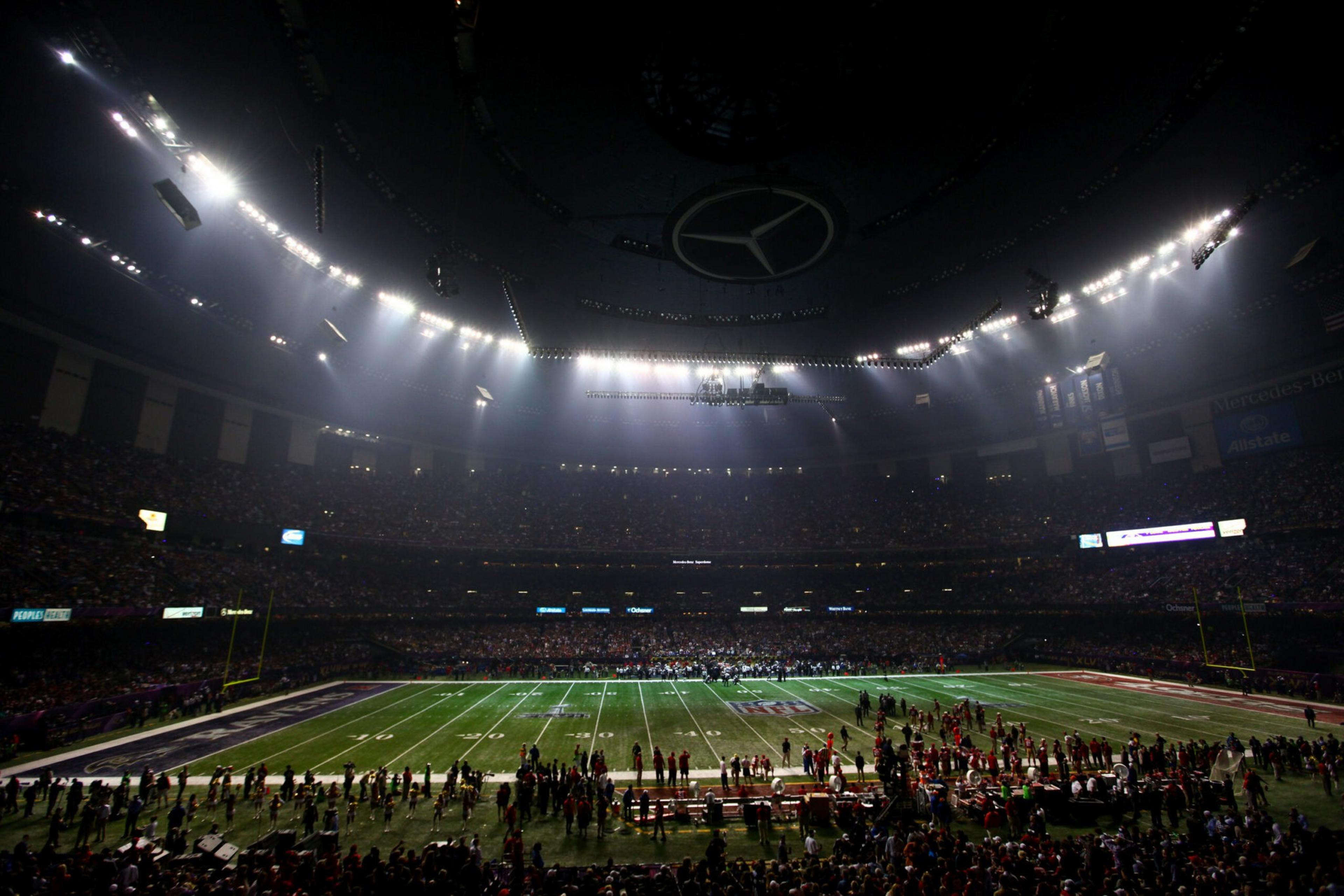 A dimly lit football stadium with the field illuminated, showing players and staff on the sidelines and a large Mercedes-Benz logo on the ceiling.