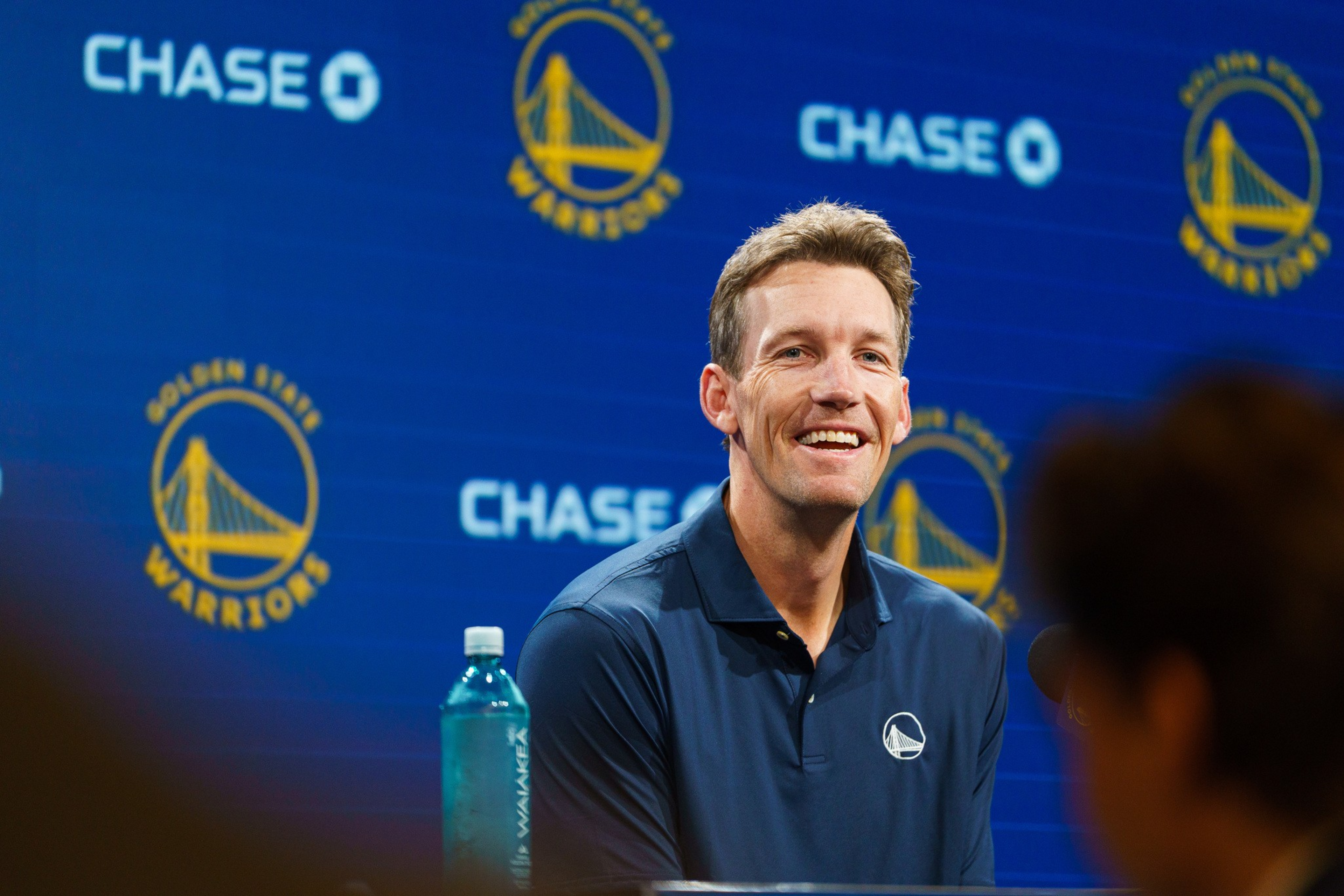 A man in a navy polo shirt with a Golden State Warriors logo smiles during a press conference, with a water bottle and team logos in the background.