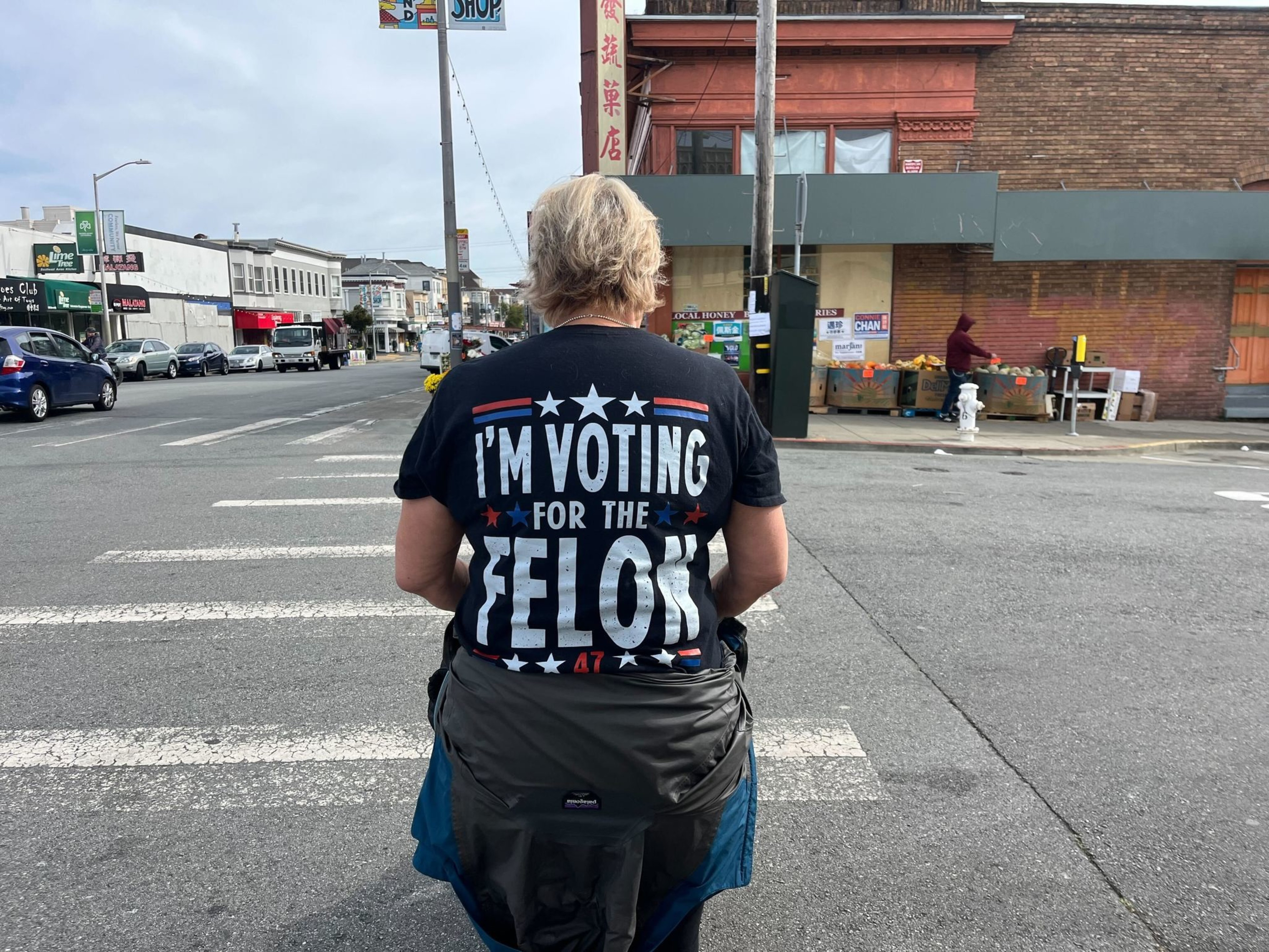 A person is standing at a crosswalk wearing a black T-shirt that says &quot;I'M VOTING FOR THE FELON&quot; in white letters. The background shows a street scene.