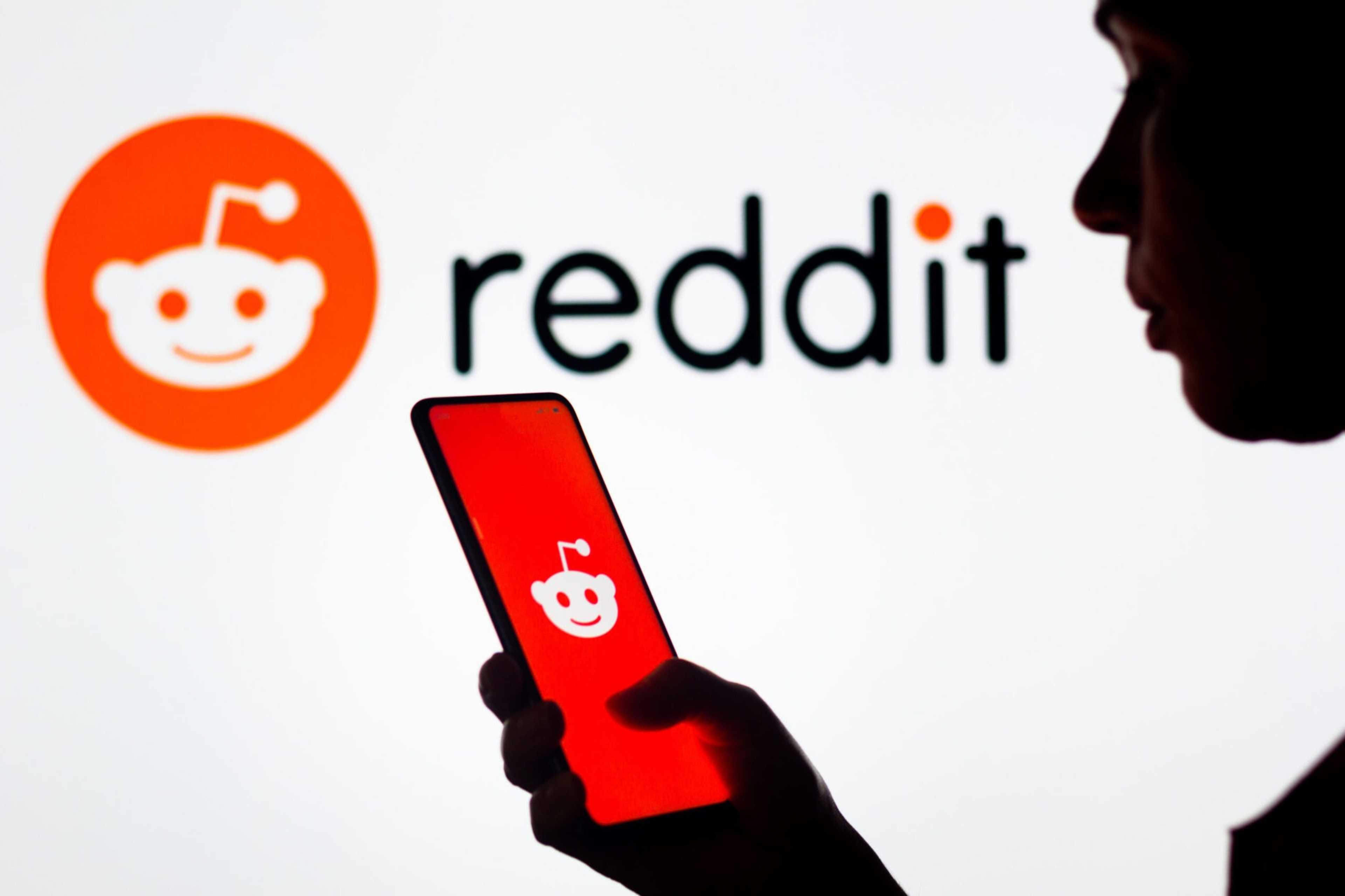 In this photo illustration, a womanâ€™s silhouette holds a smartphone with the Reddit logo displayed.