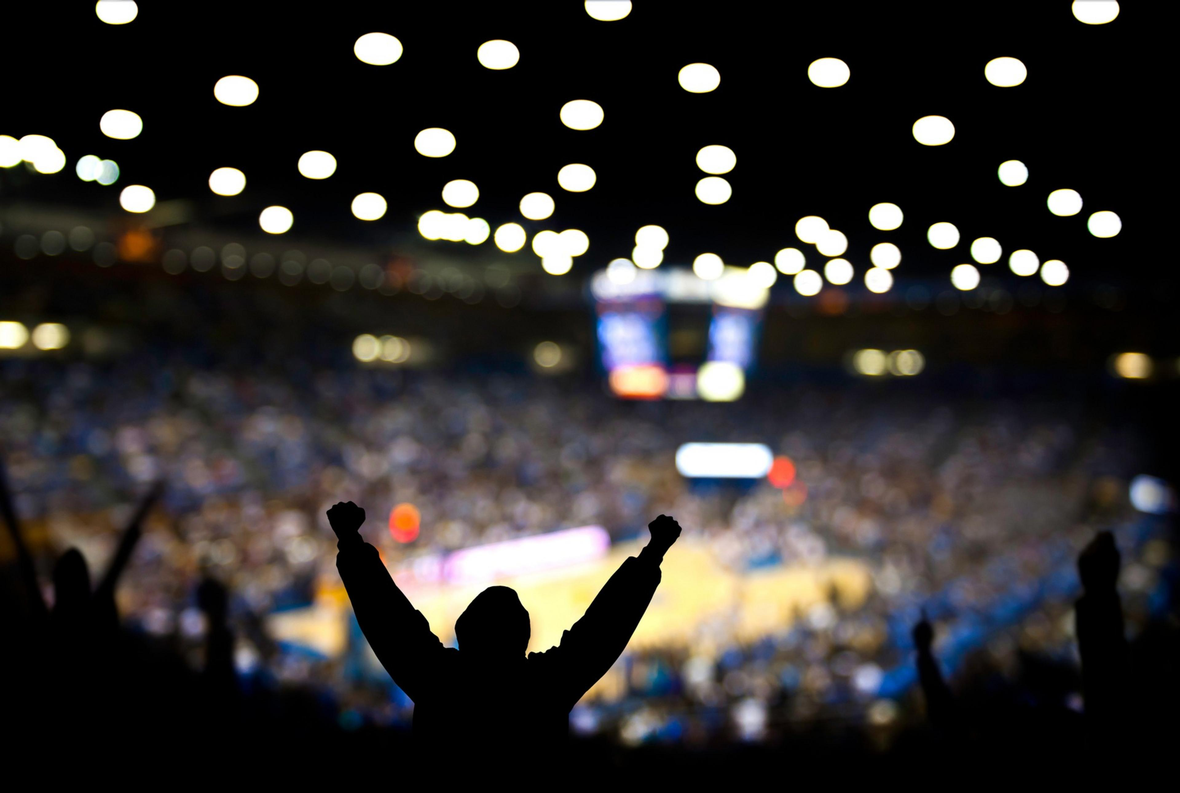 A silhouette of a cheering person is in the foreground, with a blurred, brightly lit indoor sports arena and crowd visible in the background.