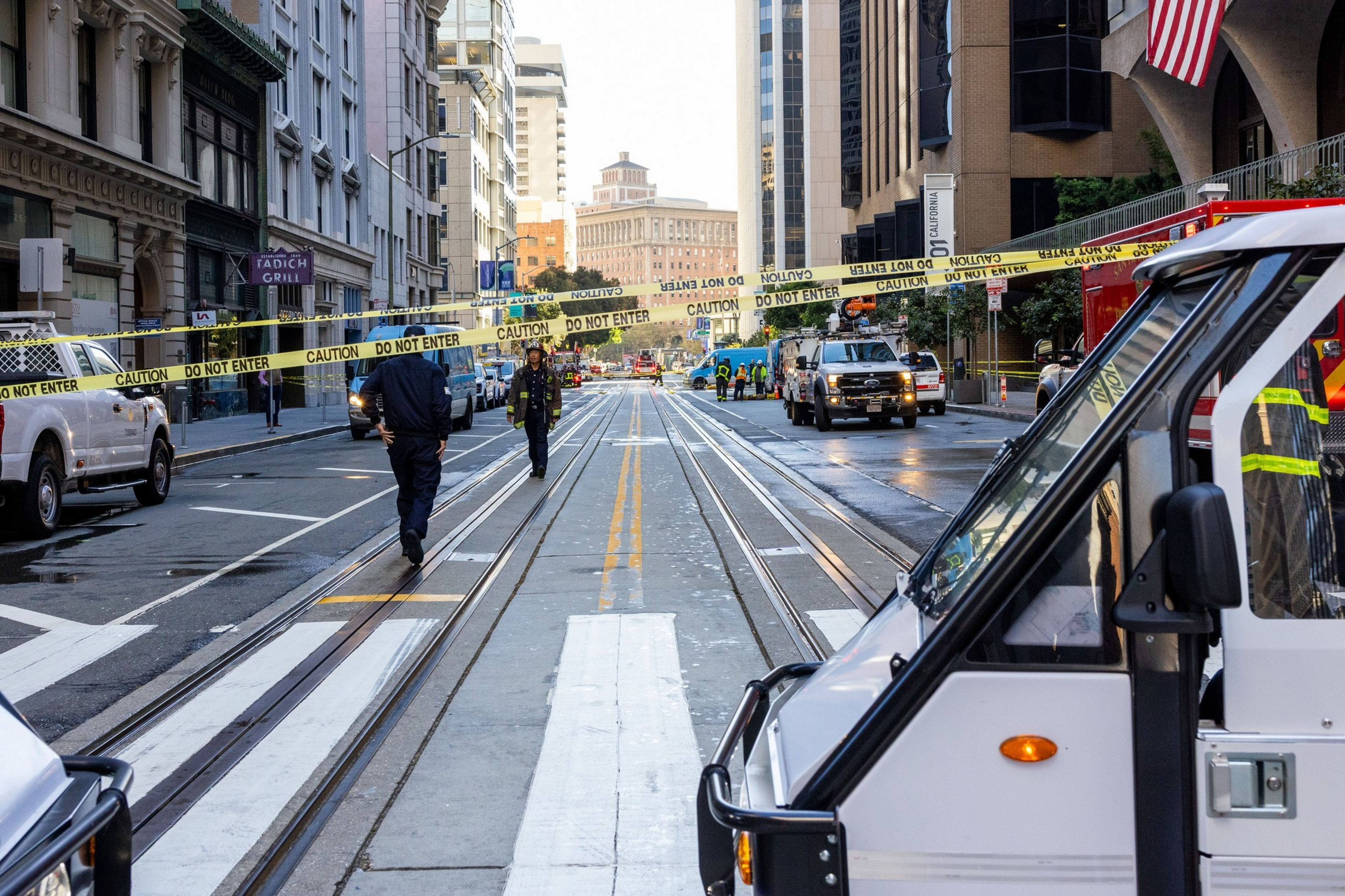 A city street is blocked off with yellow caution tape, police officers patrol the area, and vehicles are stopped near tall buildings.