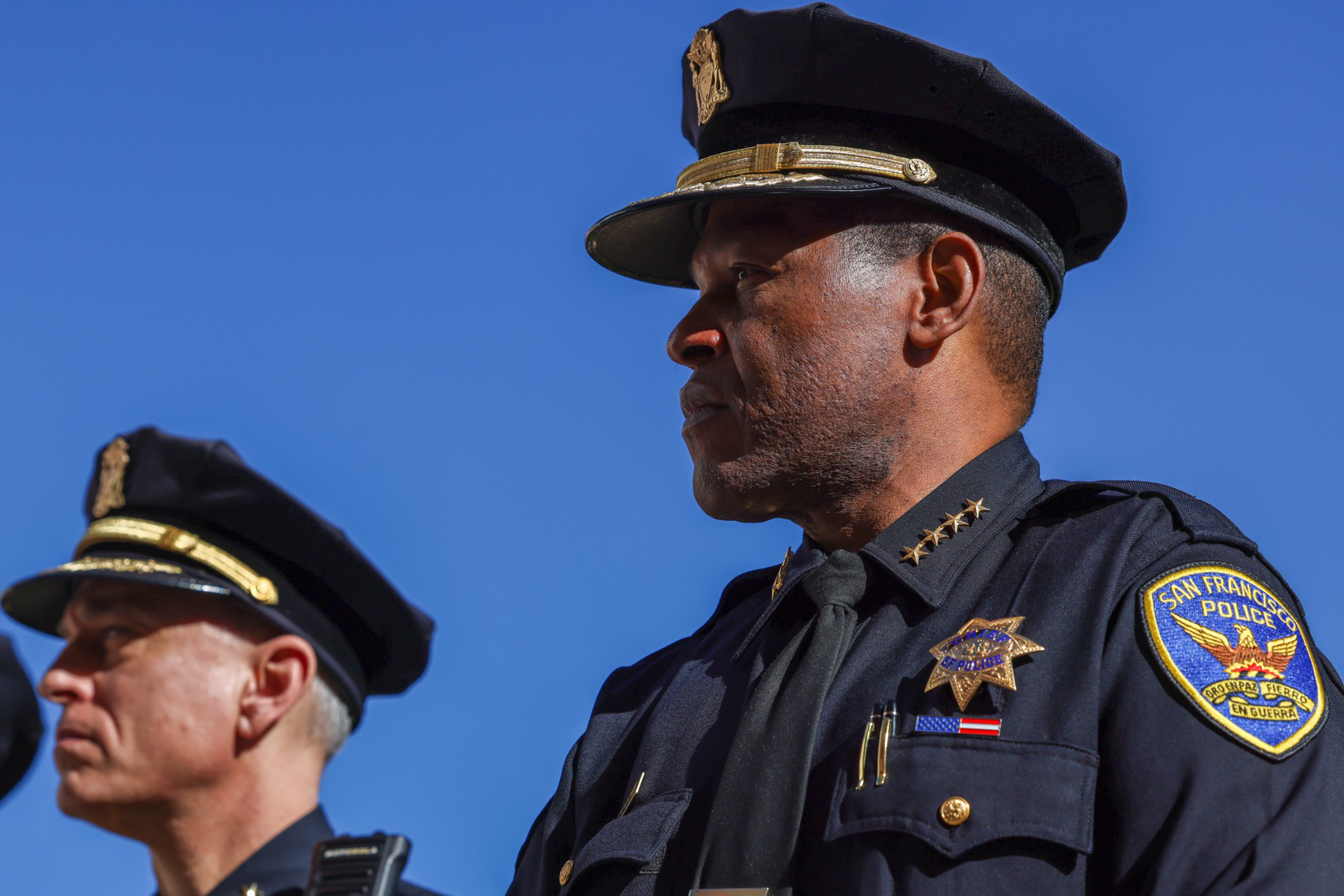 Two police officers in dark uniforms and hats stand against a clear blue sky, one with a San Francisco Police patch and four stars on his collar.