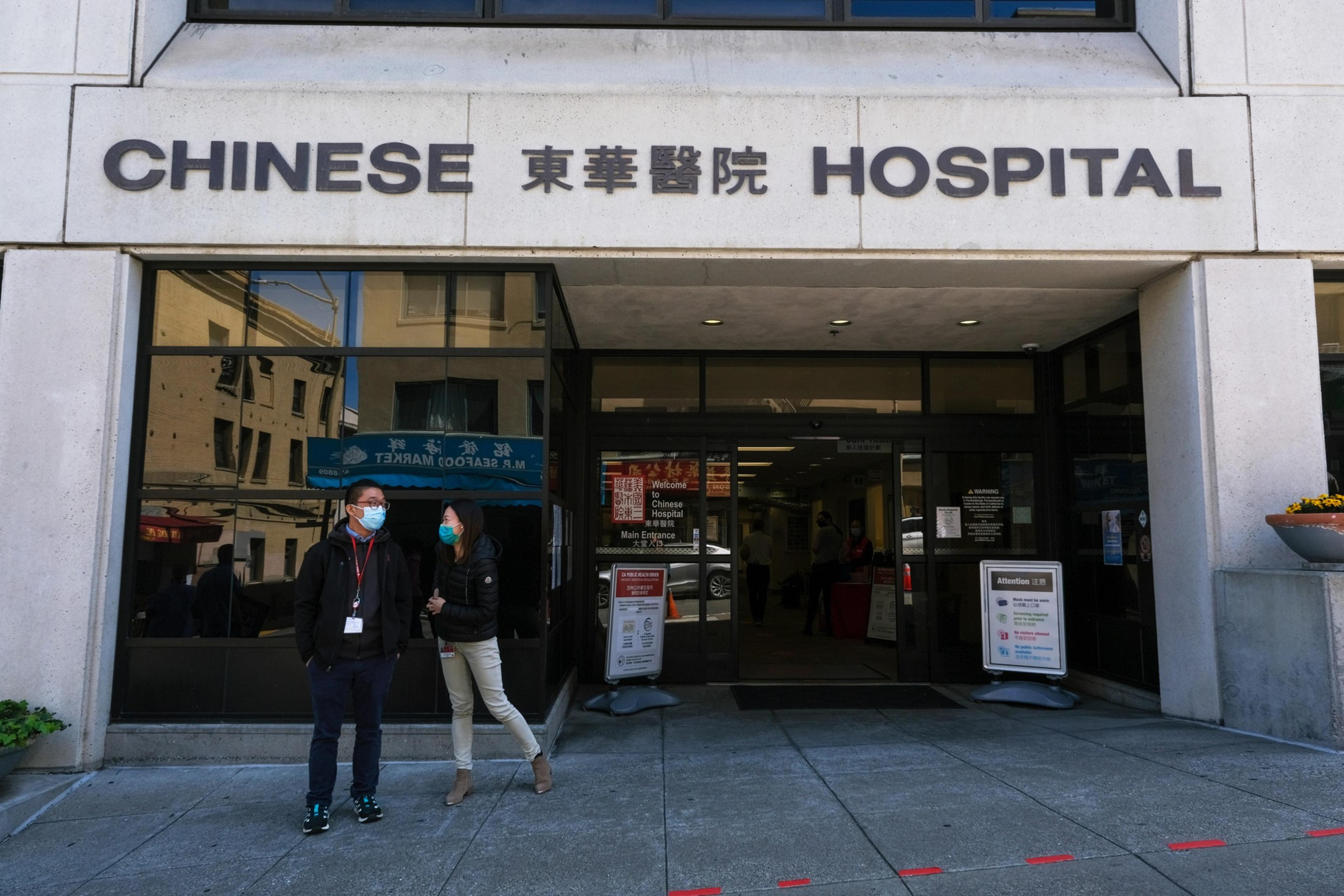 Two people wearing face masks stand outside the entrance of a building labeled "Chinese Hospital" with Chinese characters above the doorway.