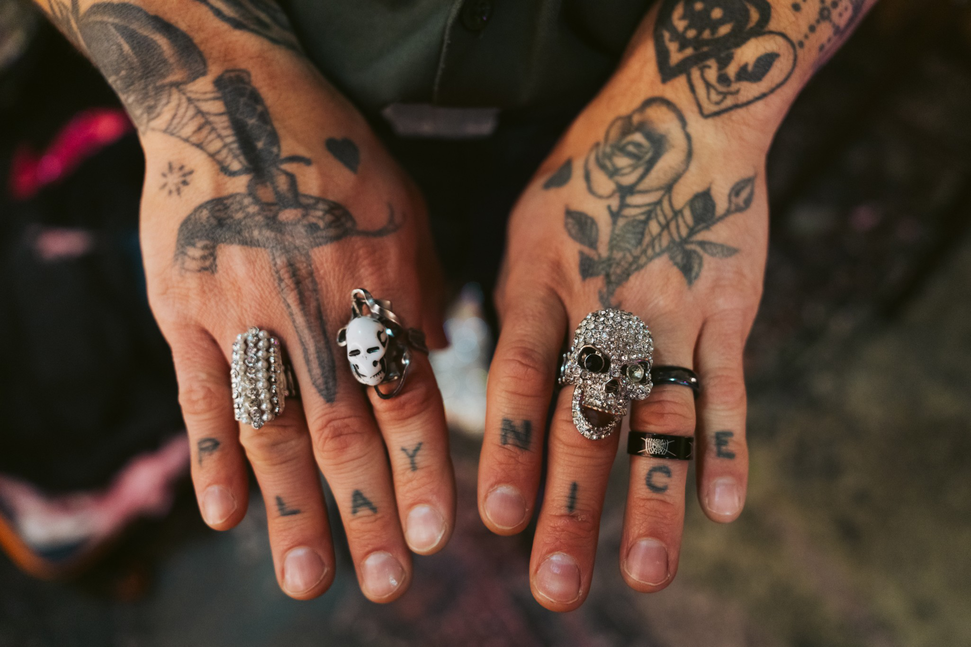 A pair of tattooed hands display elaborate rings, including a skull and panda. The fingers spell &quot;PLAY NICE&quot; with tattoos, and tattoos adorn the arms.