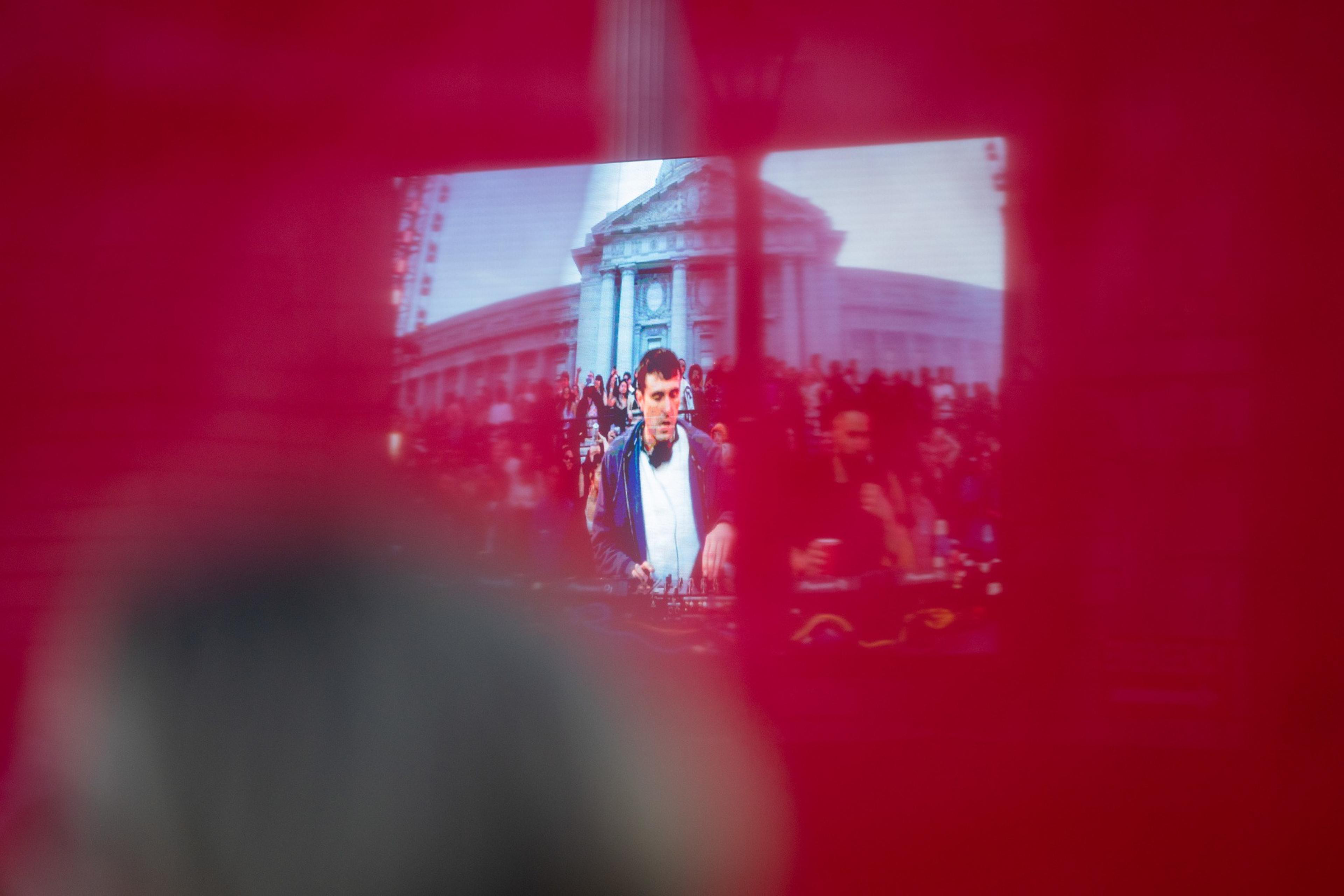 A DJ is performing with a large crowd and a grand building in the background, seen through a red filter or curtain. The DJ is focused on mixing music.