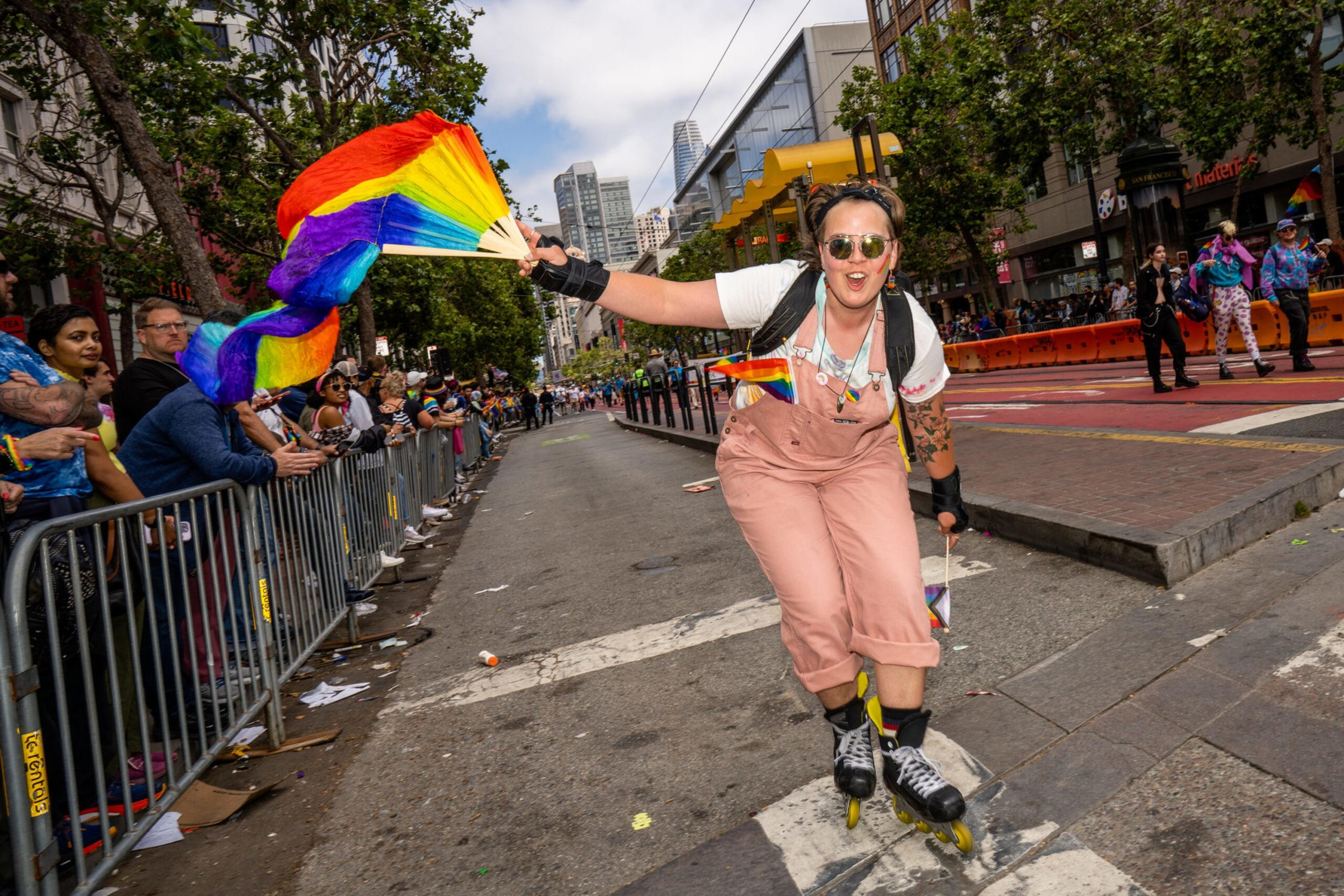 A person rollerblading down a city street waves a rainbow pride flag, wearing pink overalls and sunglasses, as a crowd watches from behind metal barricades.
