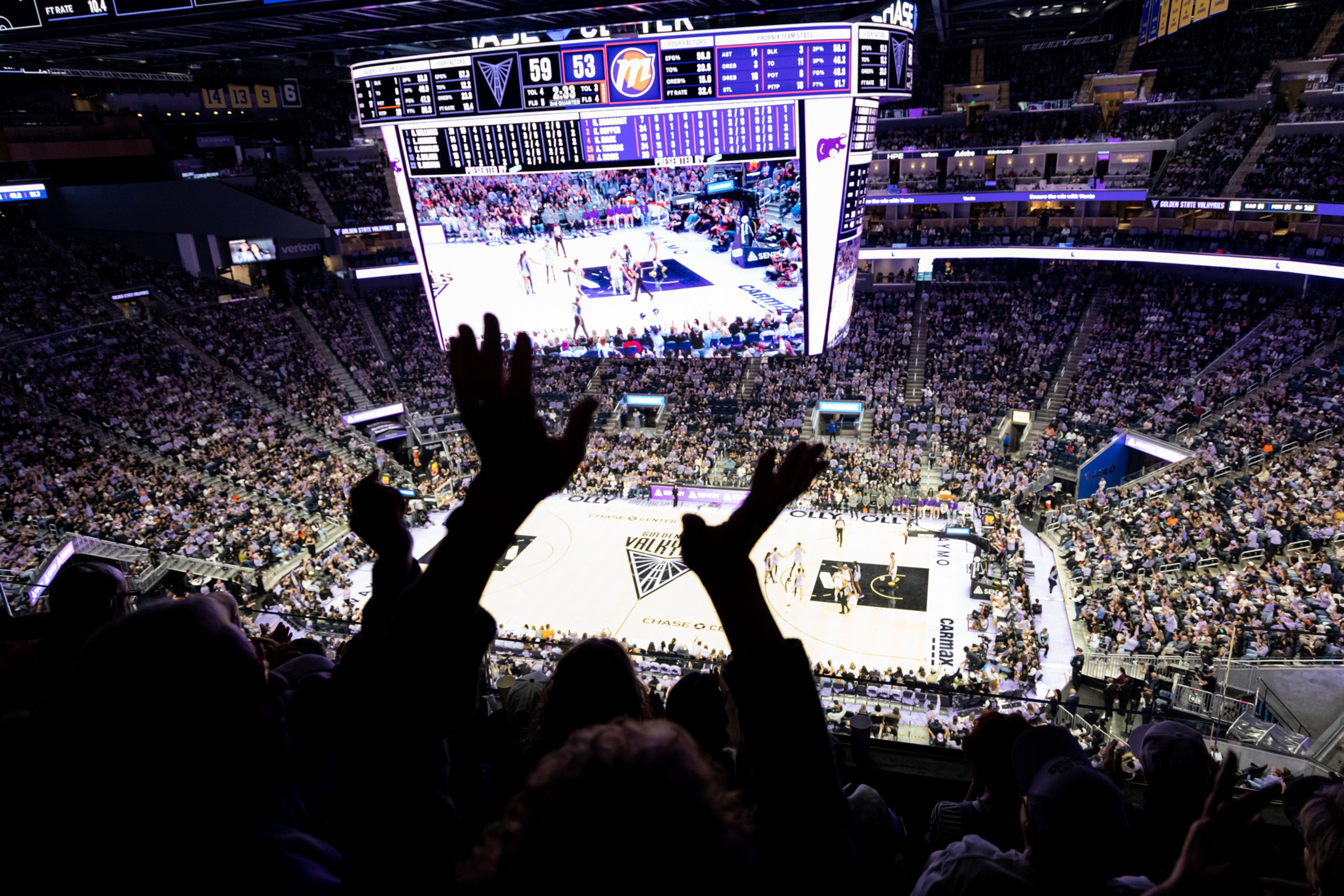 A crowded basketball arena during a game with fans raising their hands, a brightly lit court, and a large scoreboard showing the score and game details.