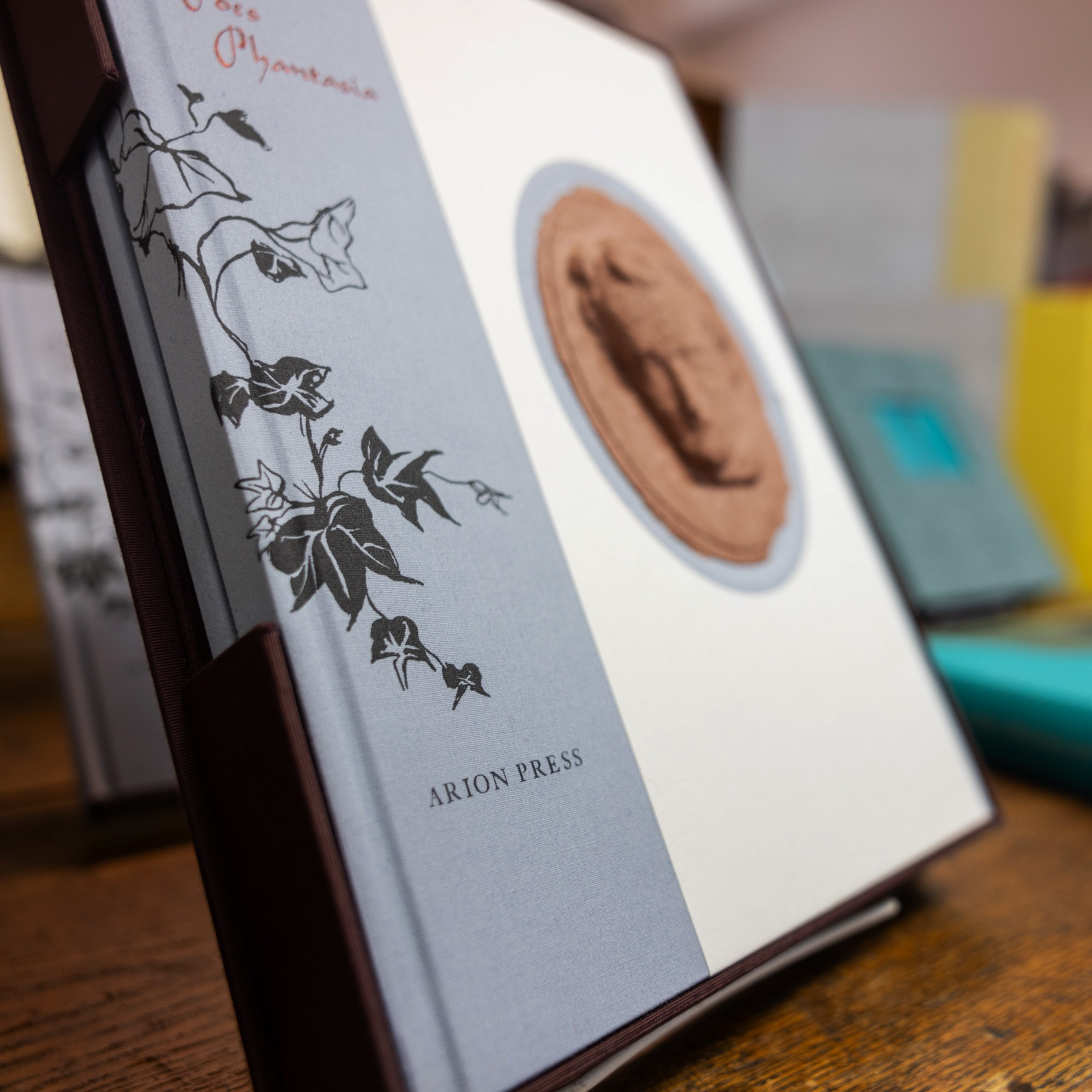 This image features an elegant book with a gray cover, adorned with a black floral design and a raised seal. The spine shows the text &quot;Arion Press.&quot;