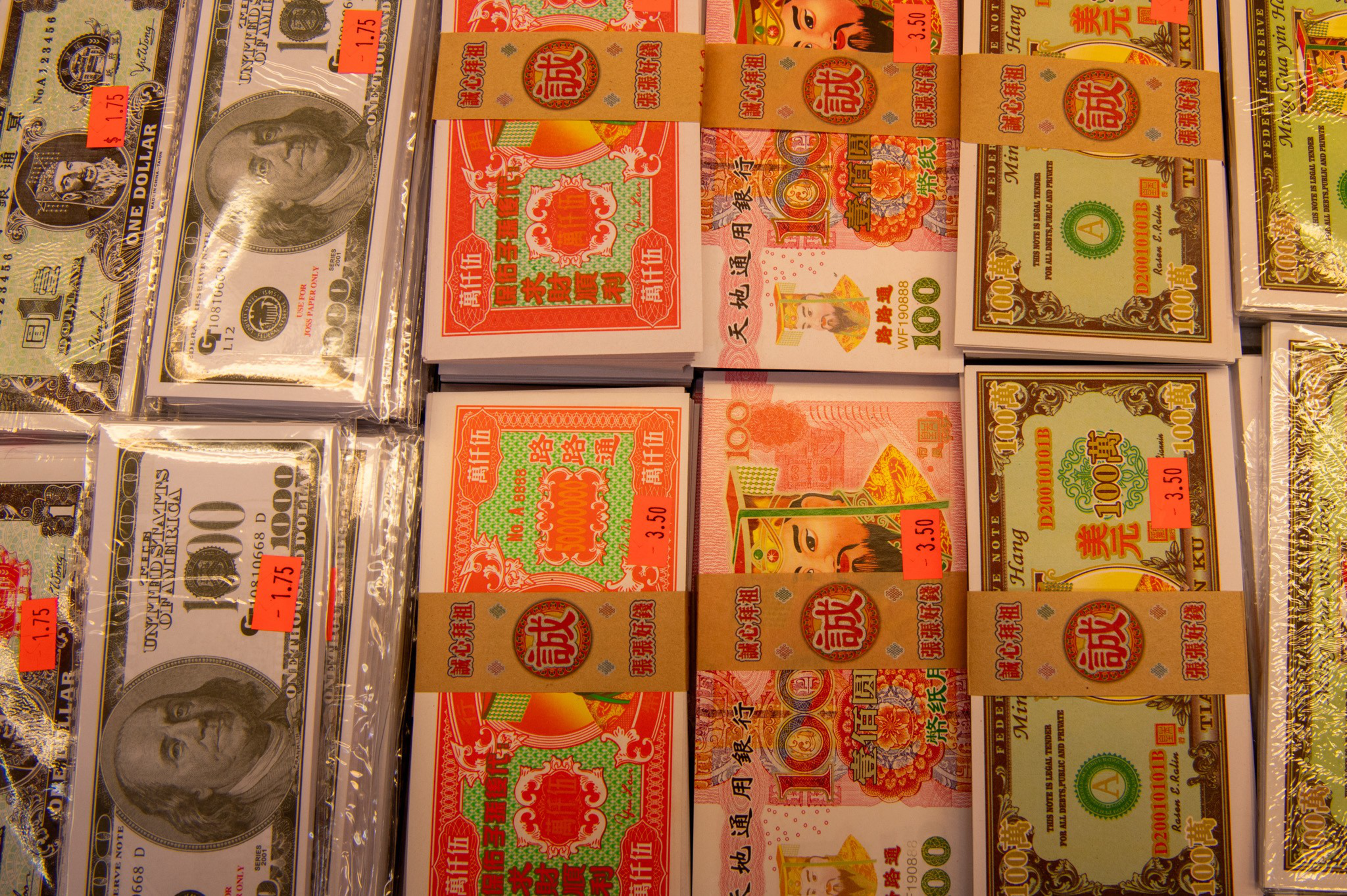 Stacks of imitation banknotes resembling US dollars and colorful Chinese joss paper bills, each with price tags attached, are displayed on a market stall.