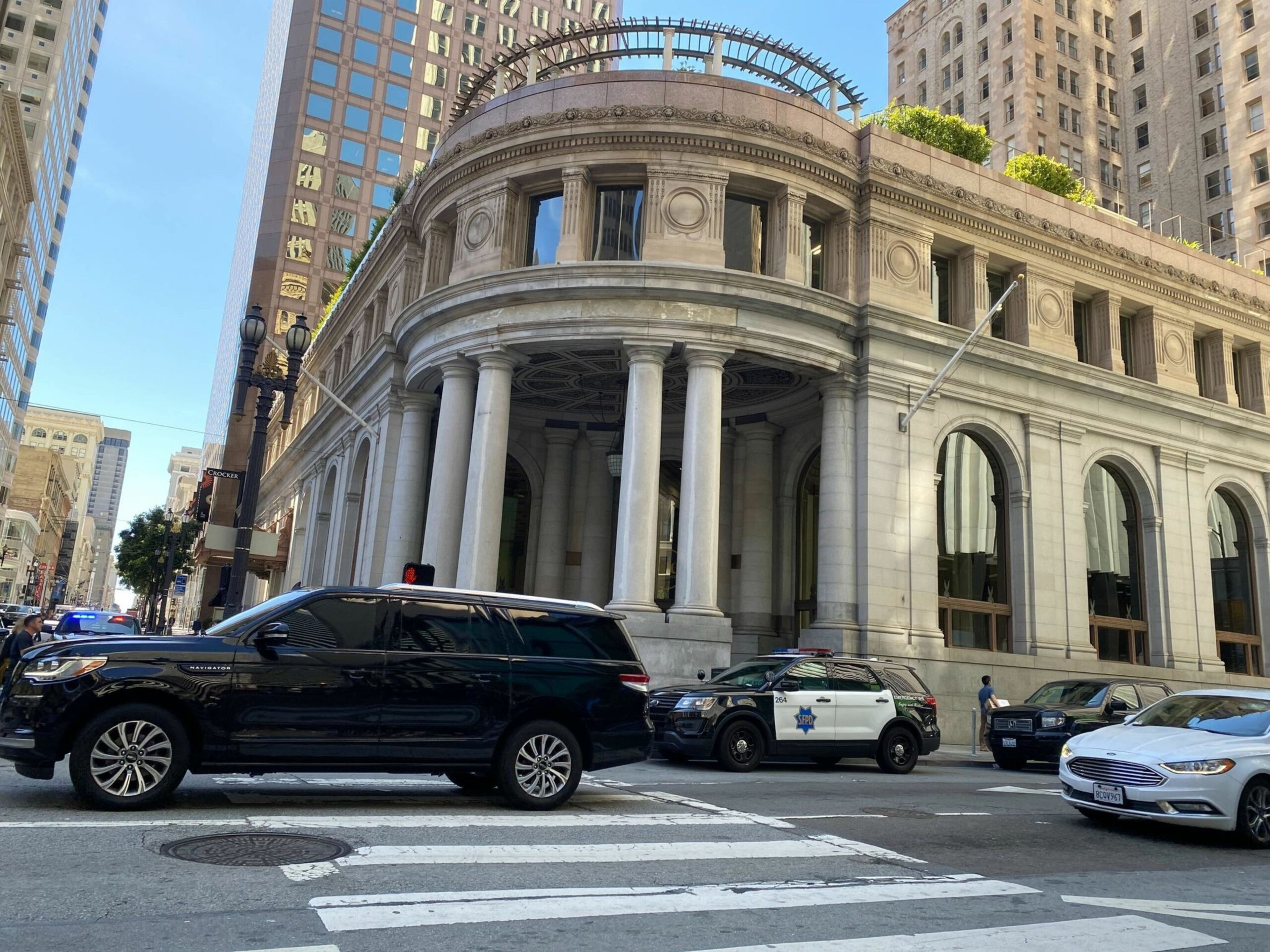 A historic stone building with tall columns and arched windows sits at a city intersection with several cars, including a police SUV, waiting at a crosswalk.