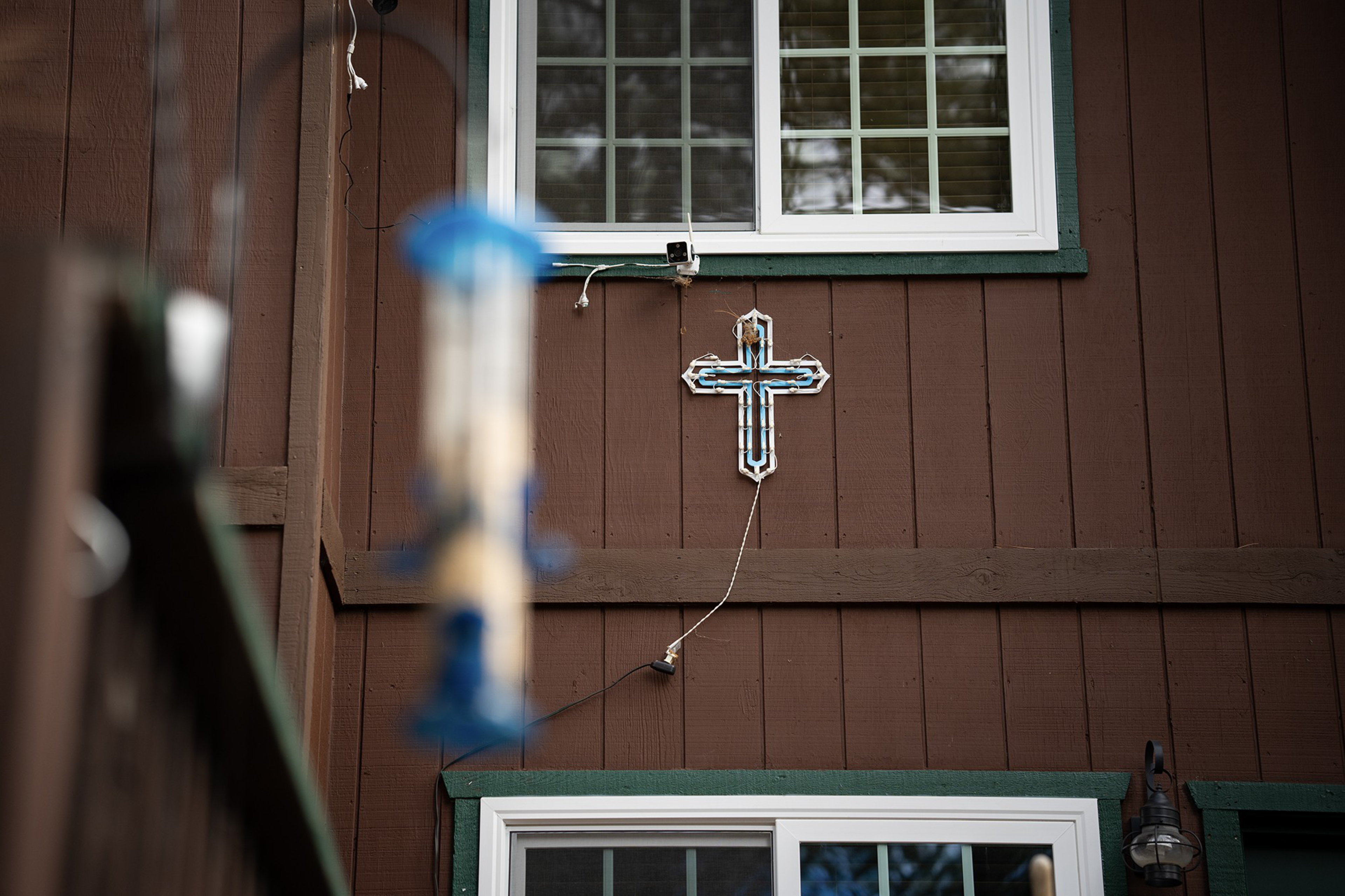 A blue and white cross light hands from the exterior of a brown house with a green trim around white windows