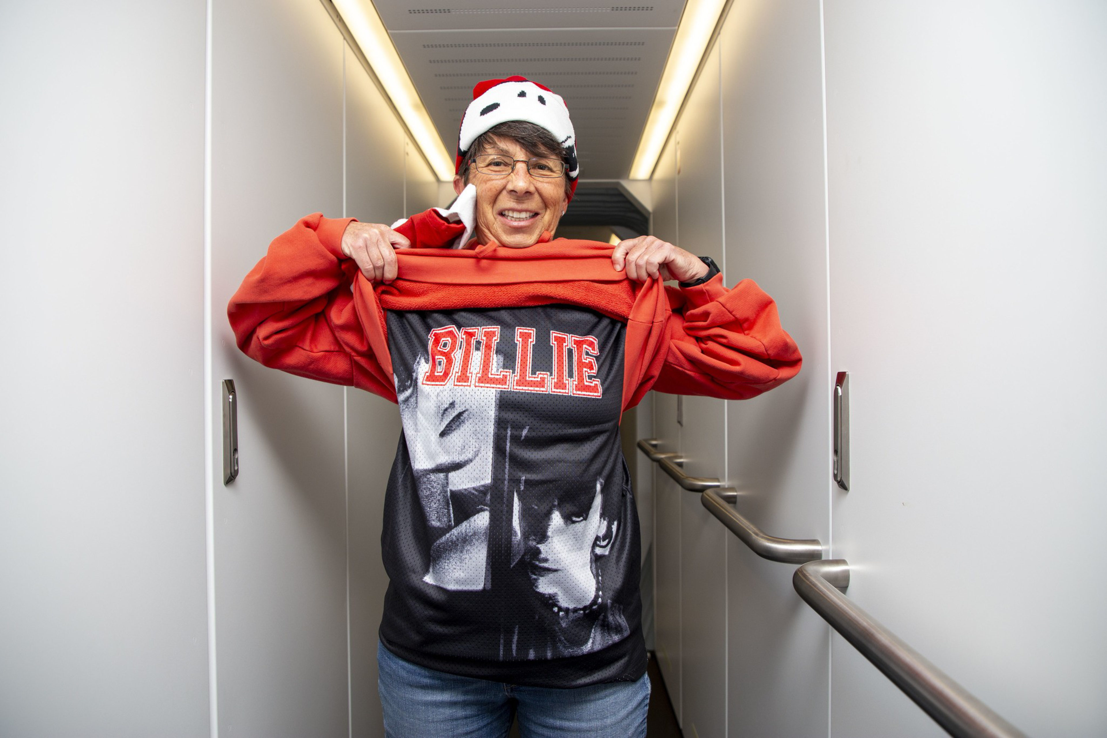 A person standing in a hallway lifts a red jacket to reveal a black shirt with &quot;BILLIE&quot; printed in red. They're wearing a Santa hat and glasses.