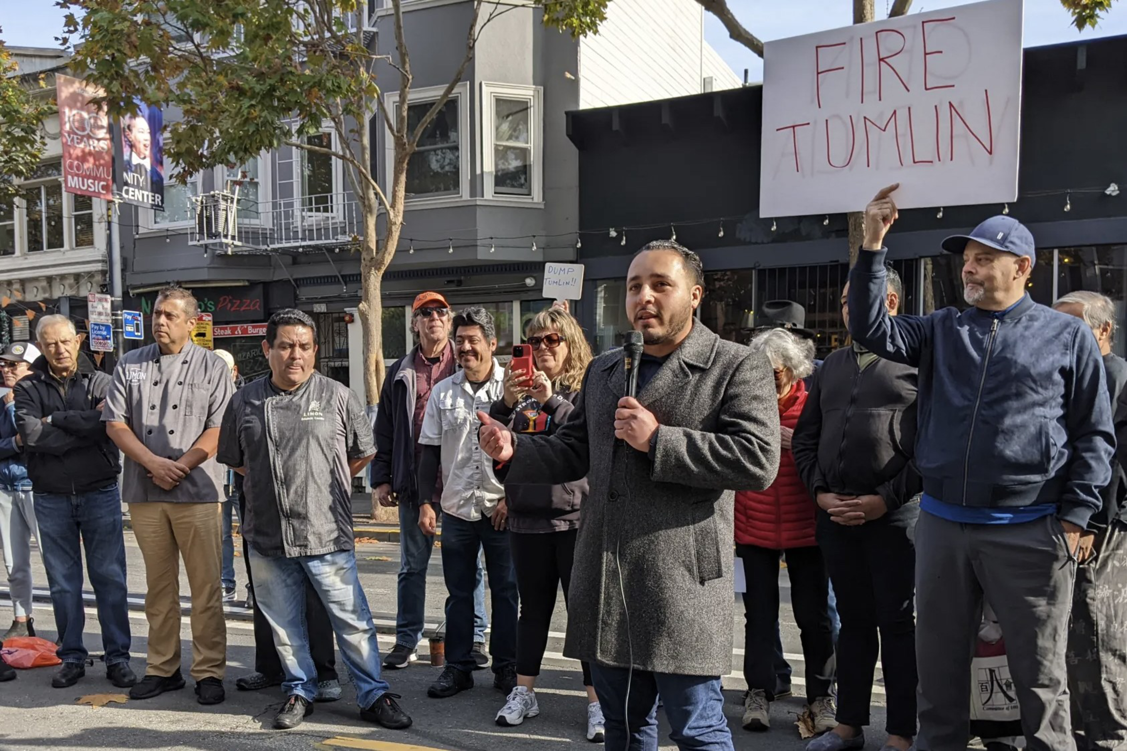 a crowd of people in a street stands behind someone holding a microphone