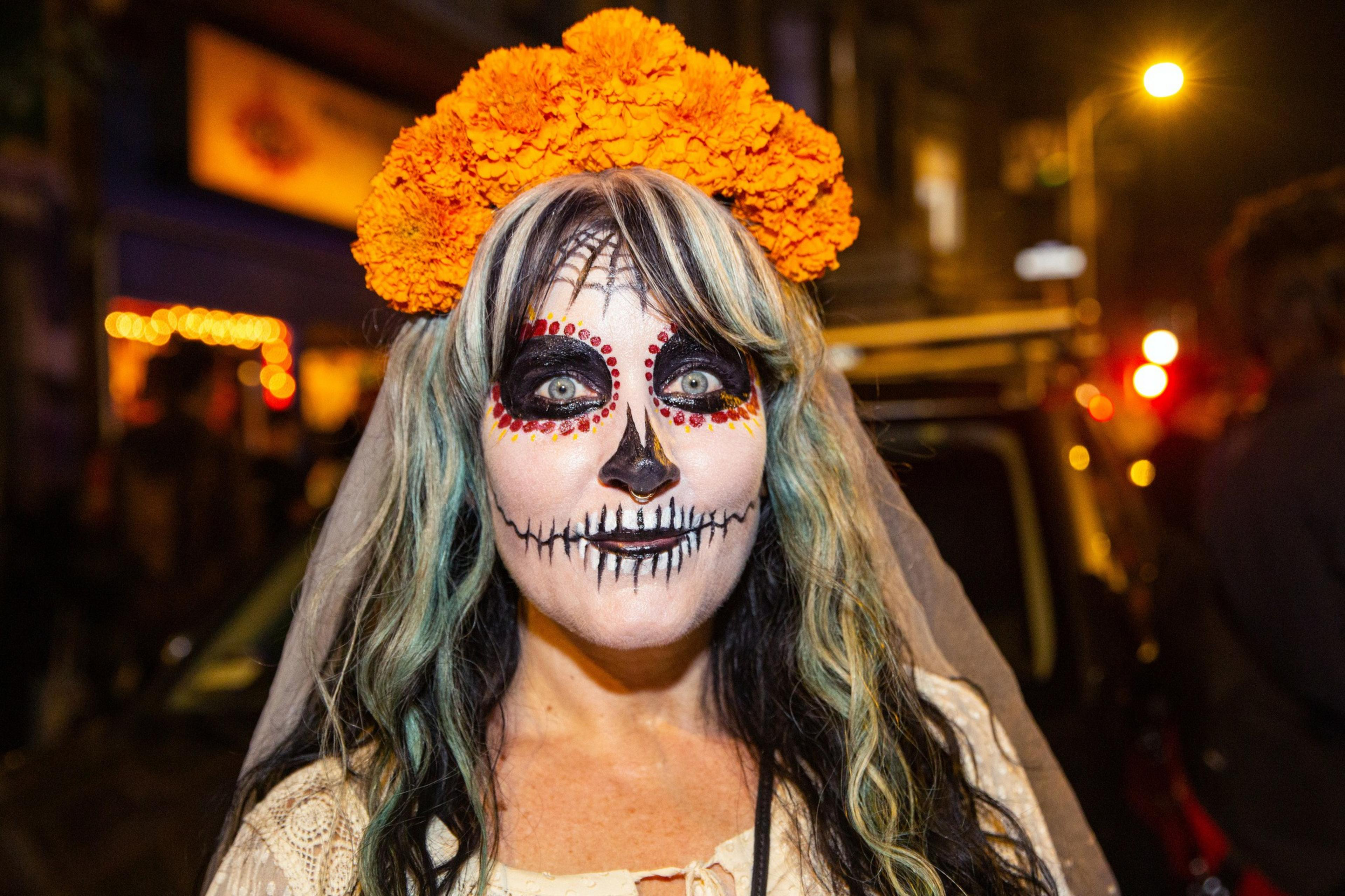 A person with Day of the Dead face paint has black and white skeletal makeup with red dots, grayish hair streaks, and an orange marigold flower crown.