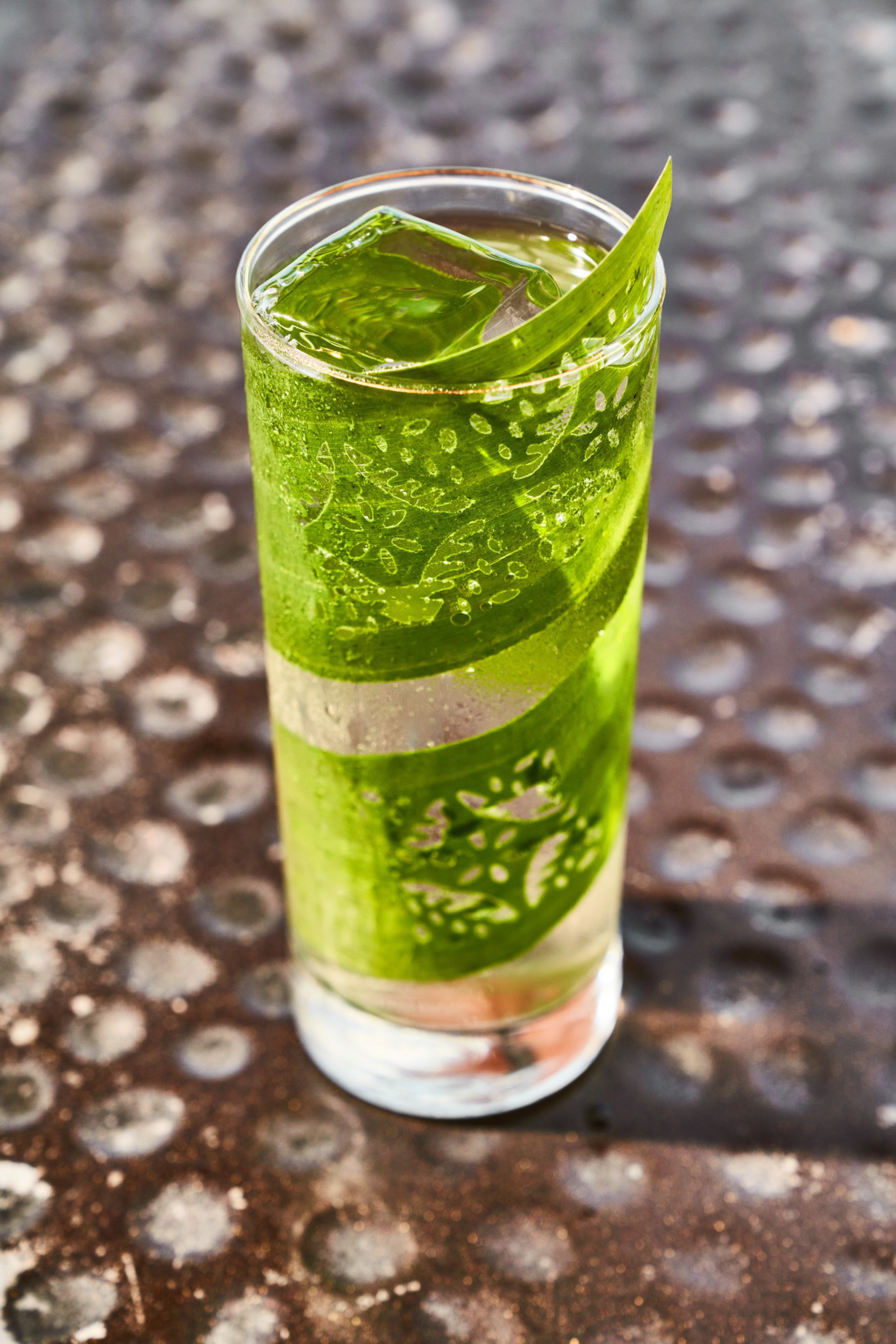 A tall glass contains a green drink with ice and a leafy garnish, set on a textured surface with a pattern of circular marks.