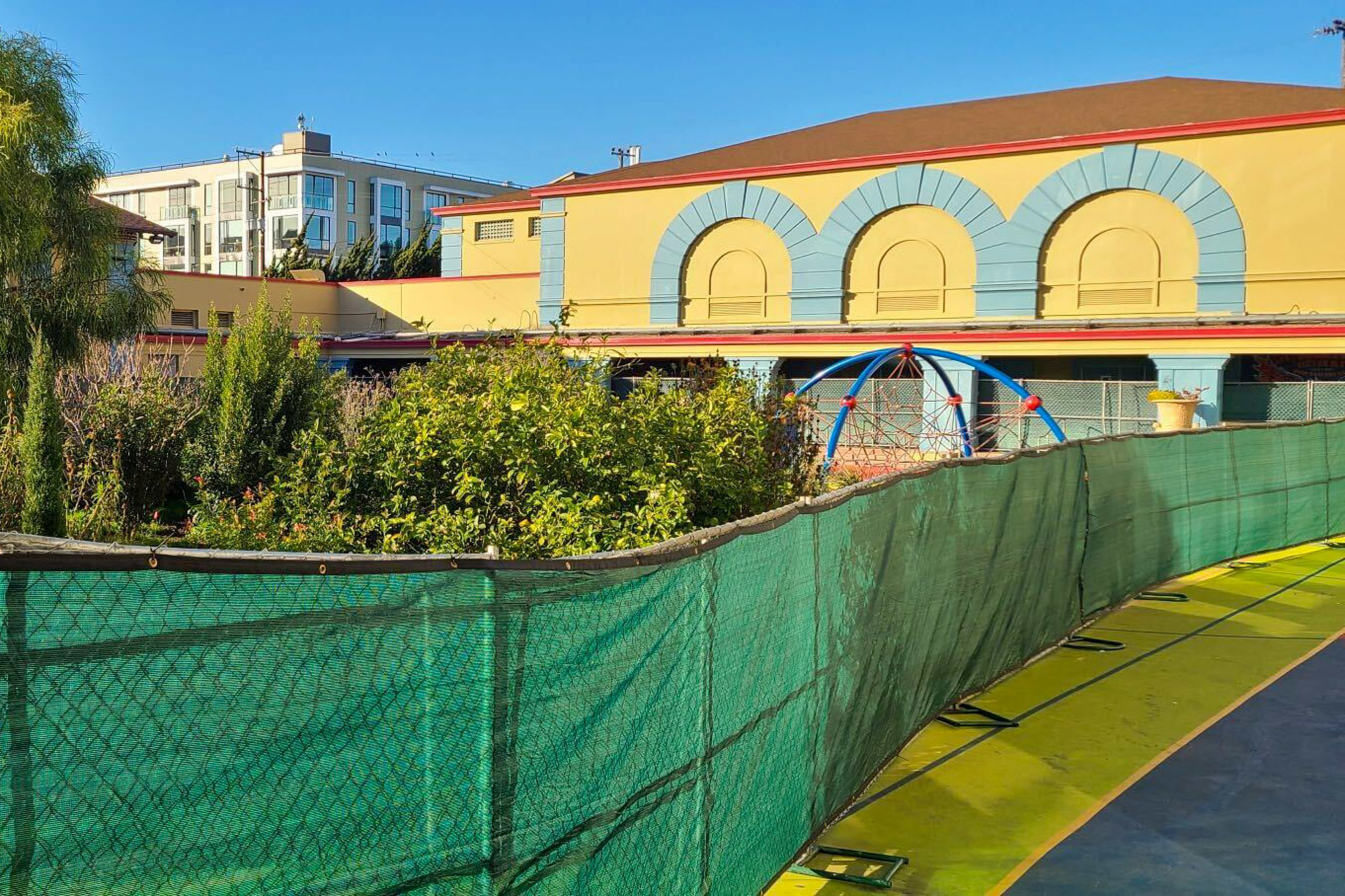 A building with yellow walls and blue arches stands behind a green mesh fence, with tall bushes and a blue and red climbing dome visible behind it.