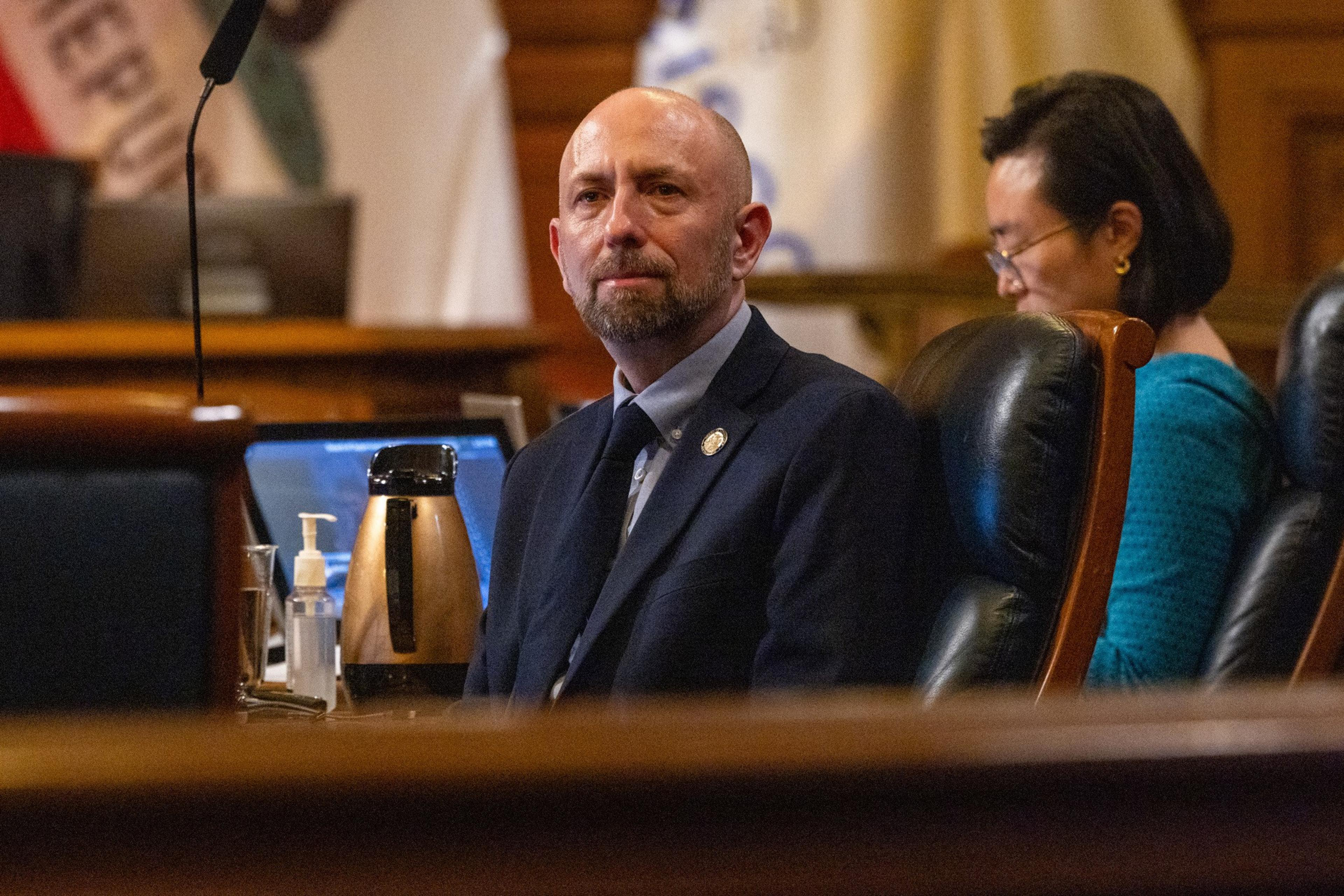 A man in a suit and tie sits in a leather chair, with a woman in blue behind him, inside a formal room with wooden furniture and flags.