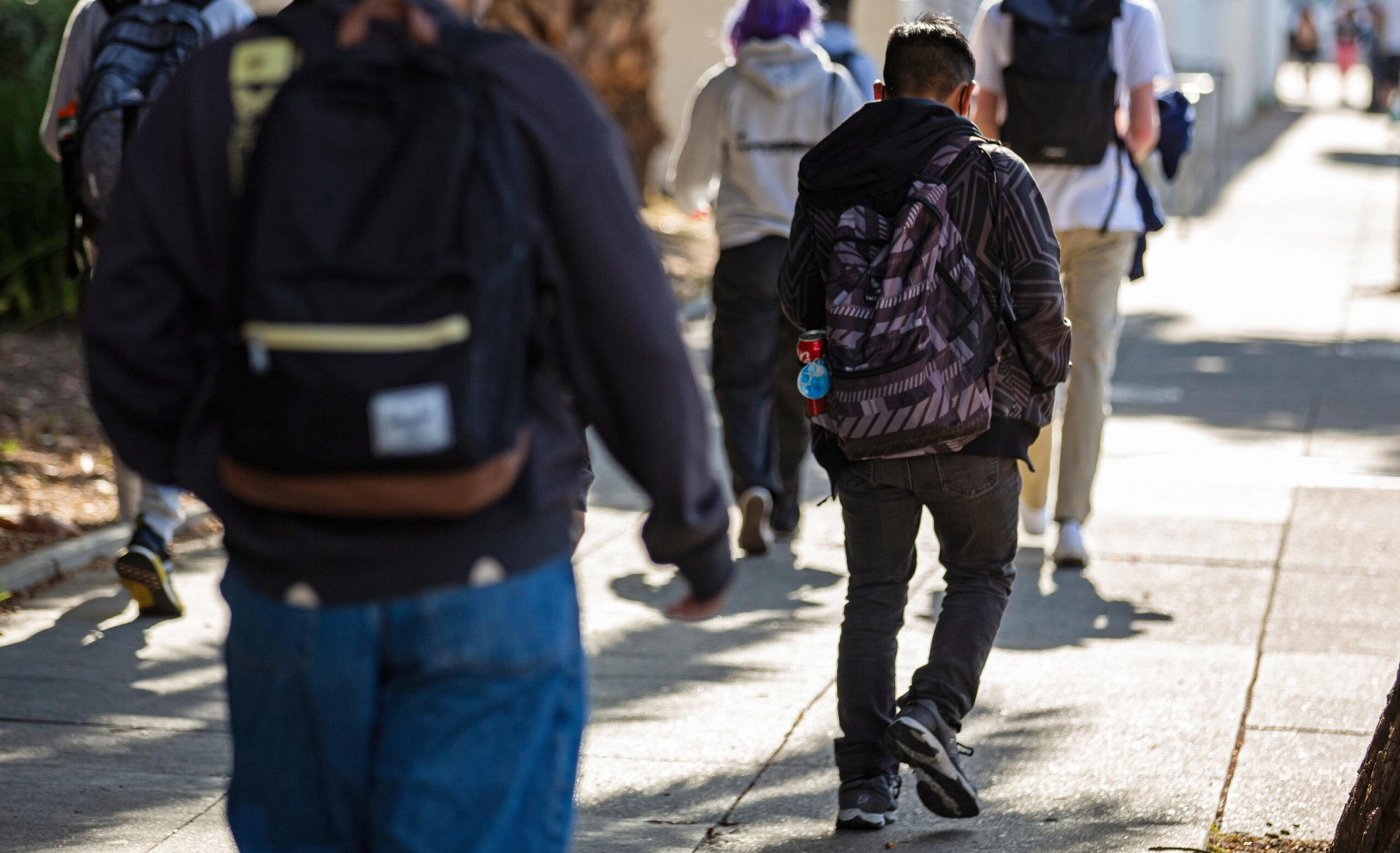 Several people wearing backpacks walk along a sunlit sidewalk, dressed casually with jackets and jeans.