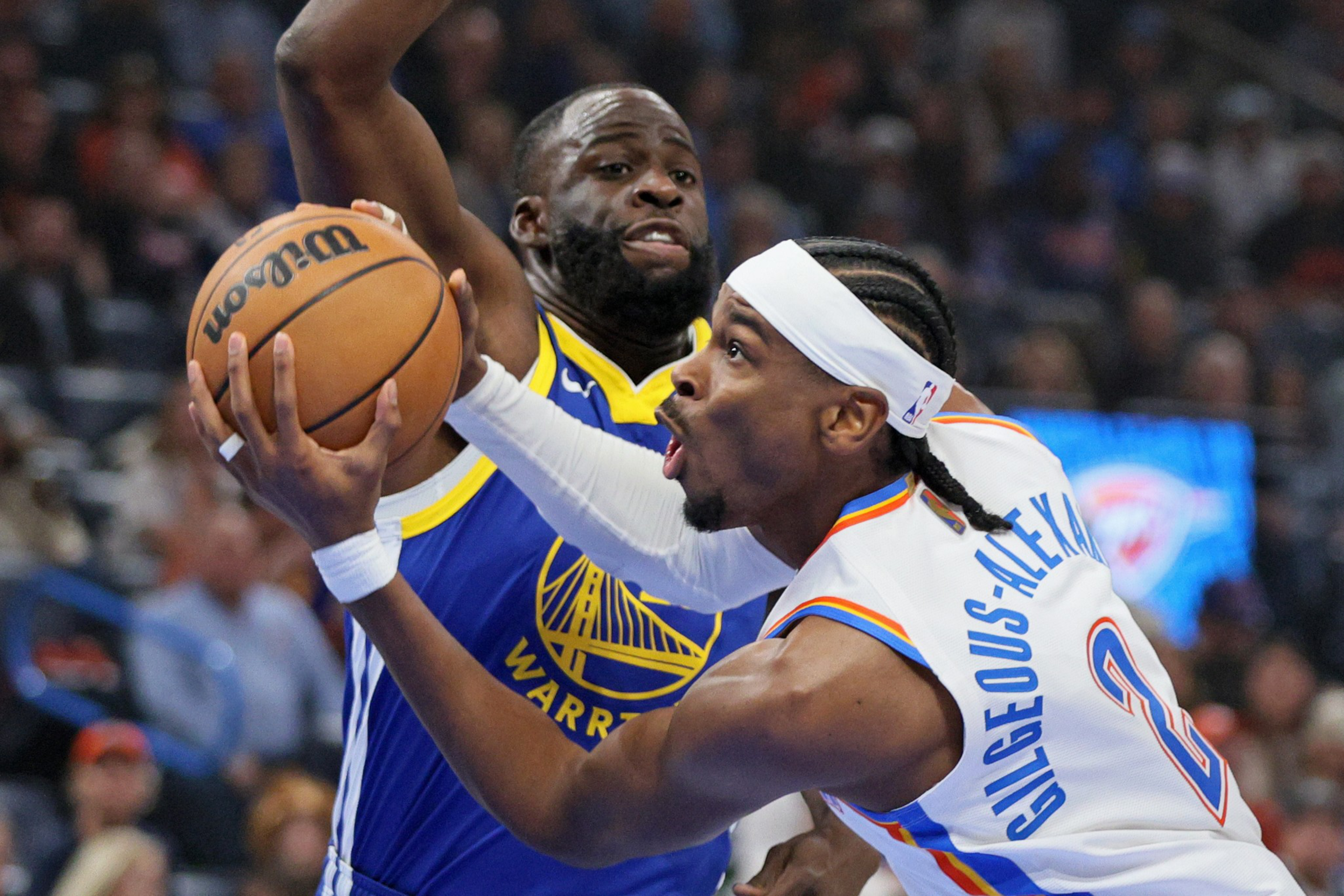 A basketball player in a white jersey attempts a shot while closely guarded by a defender in a blue and yellow Warriors uniform.