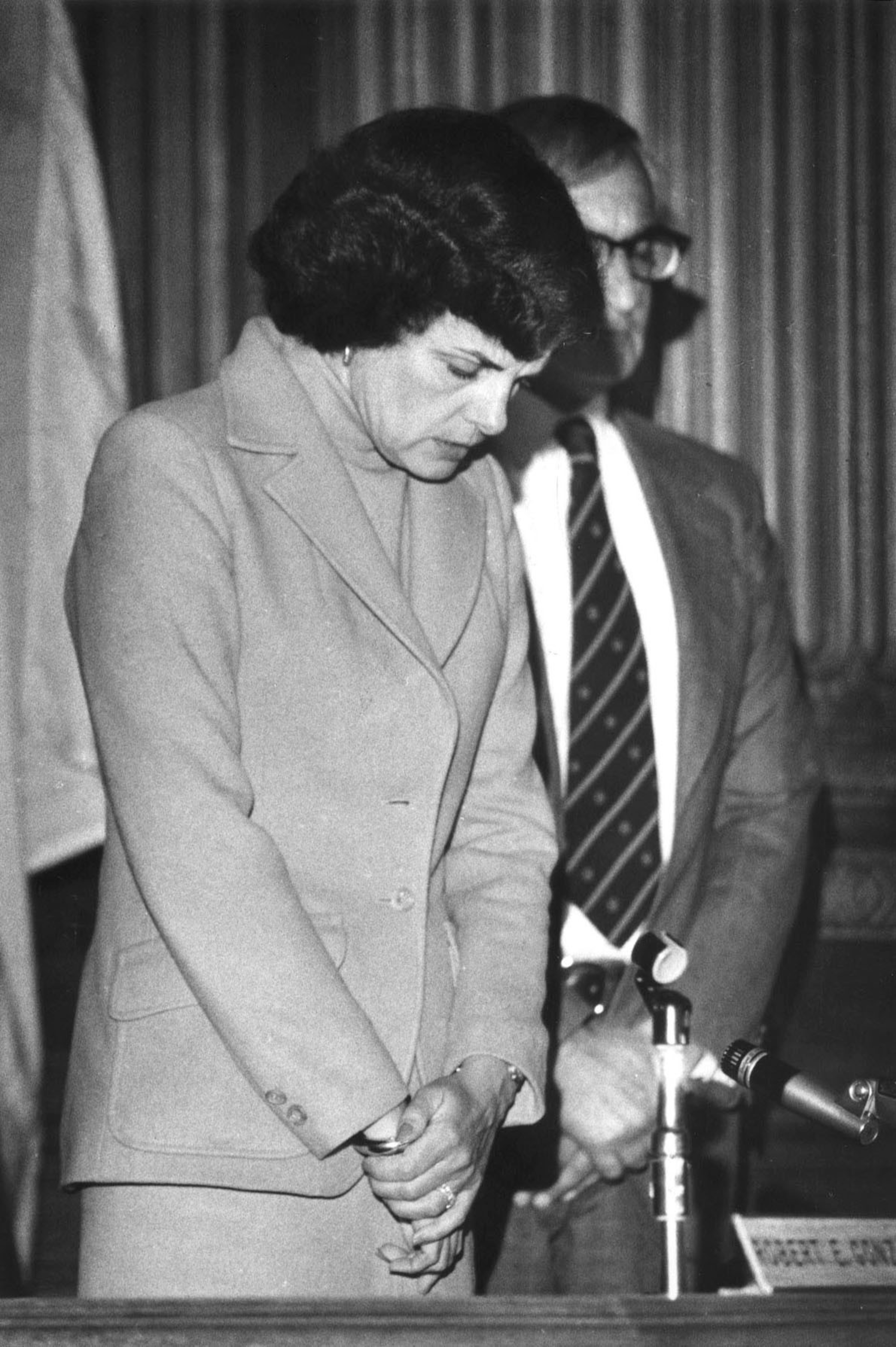 A woman in a suit stands with her head bowed and hands clasped, while a man behind her looks forward near a microphone and nameplate.