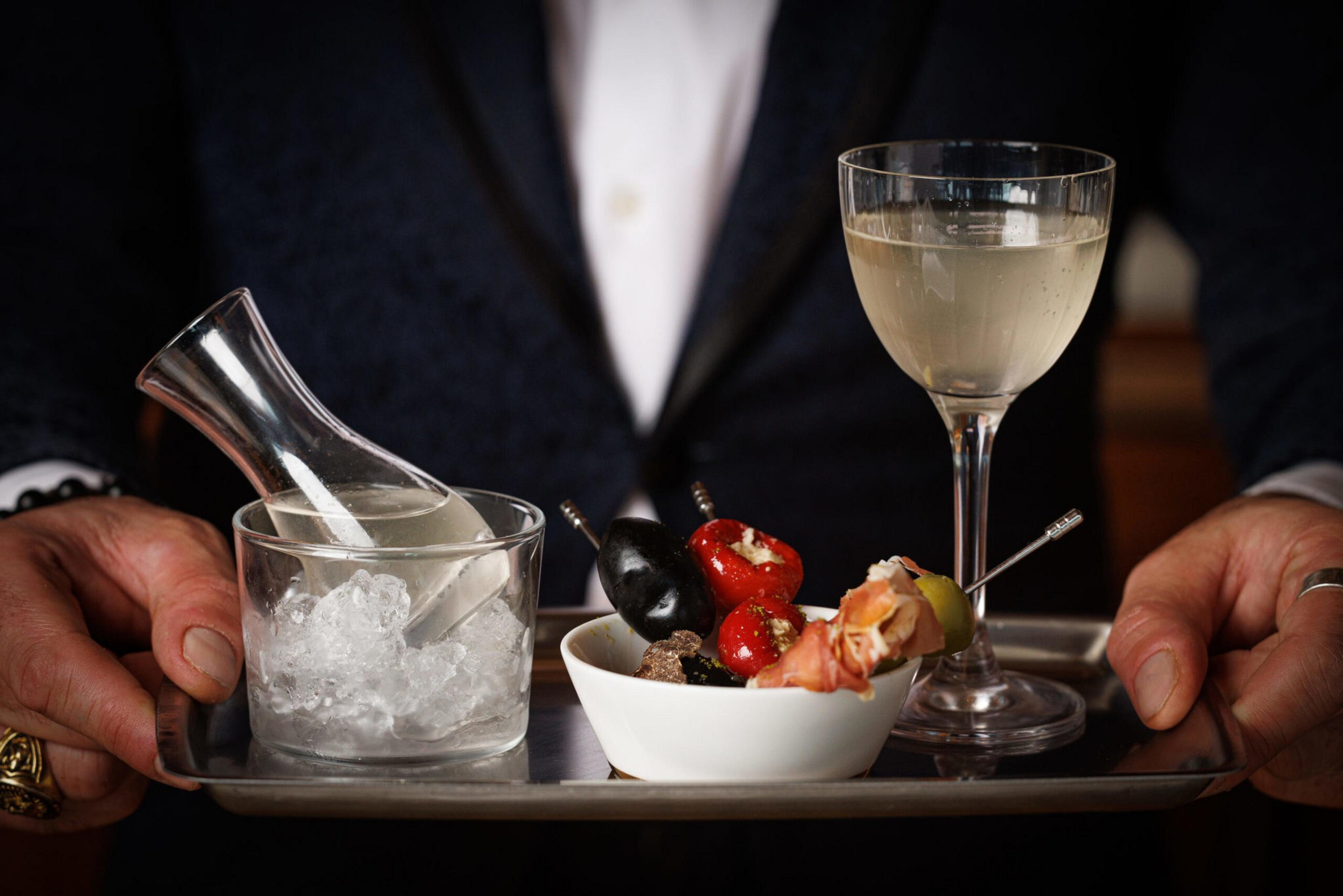 A person holds a tray with a glass of white wine, a small bowl of stuffed olives and peppers, and a carafe chilled in an ice-filled glass.