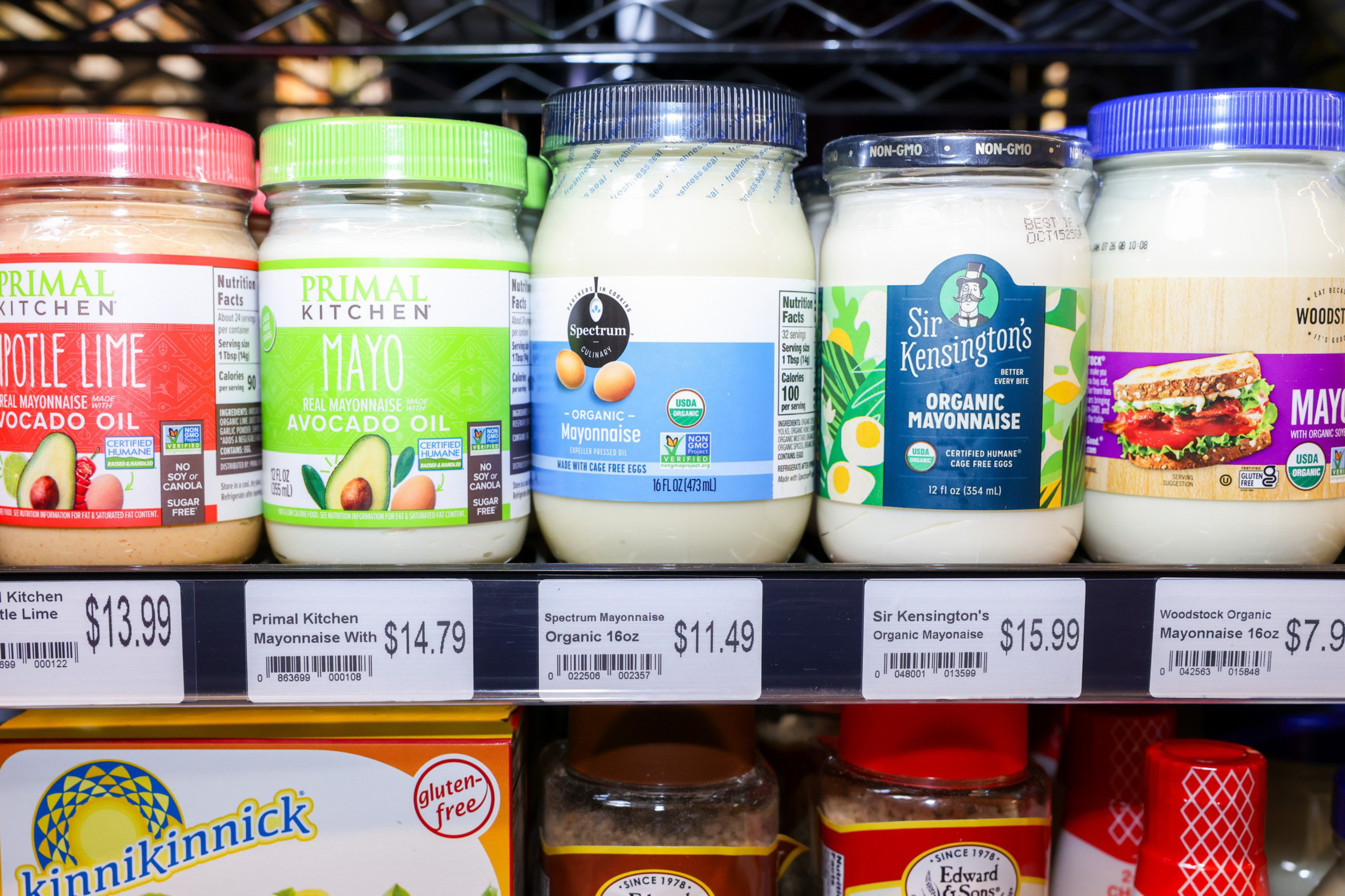 Five jars of mayonnaise are on a shelf, each with different labels and prices. They include Primal Kitchen, Spectrum, Sir Kensington's, and Woodstock brands.