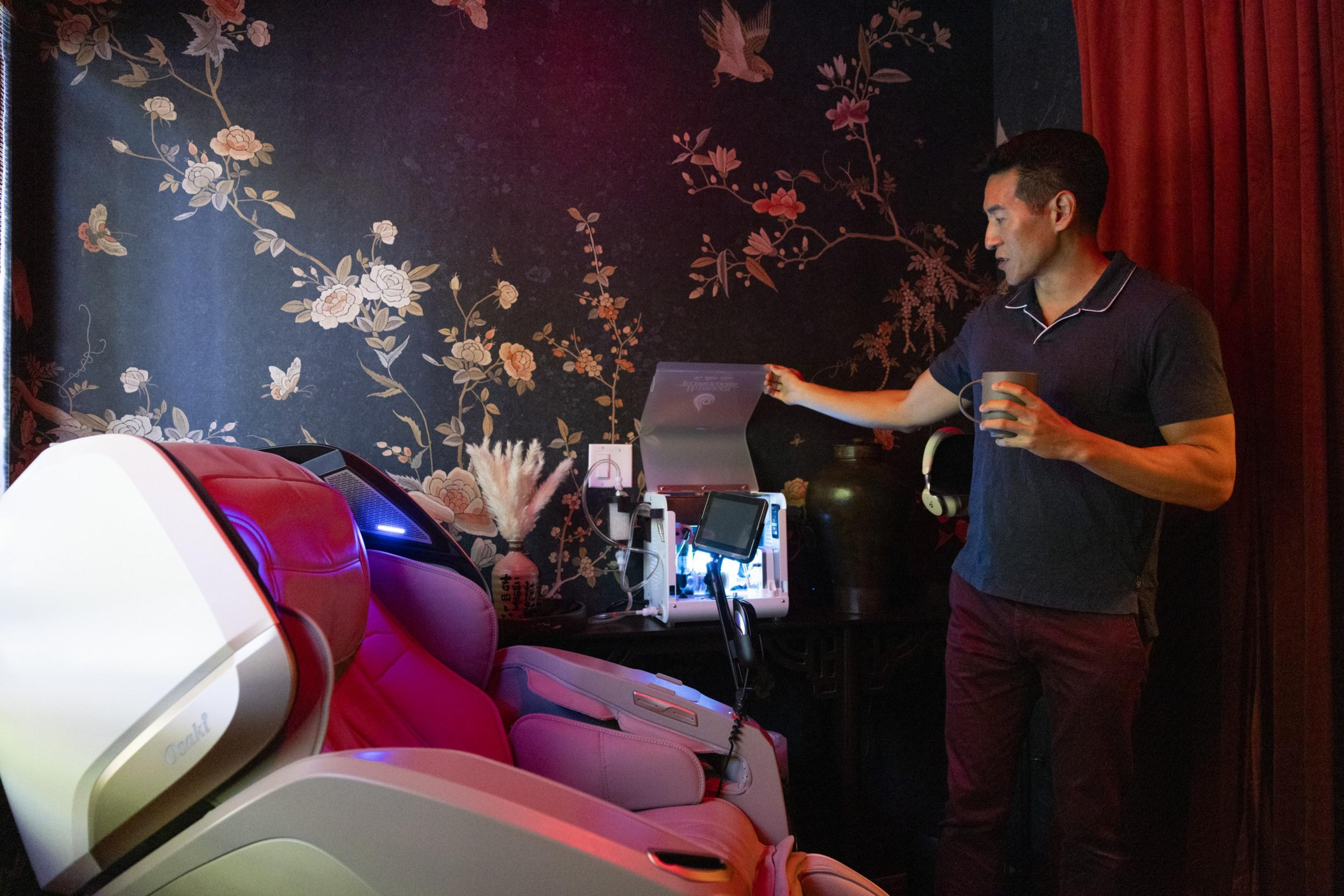 A man stands beside a futuristic, red-lit massage chair, holding a cup, with a floral wallpaper and various gadgets on a table behind him in a dimly lit room.