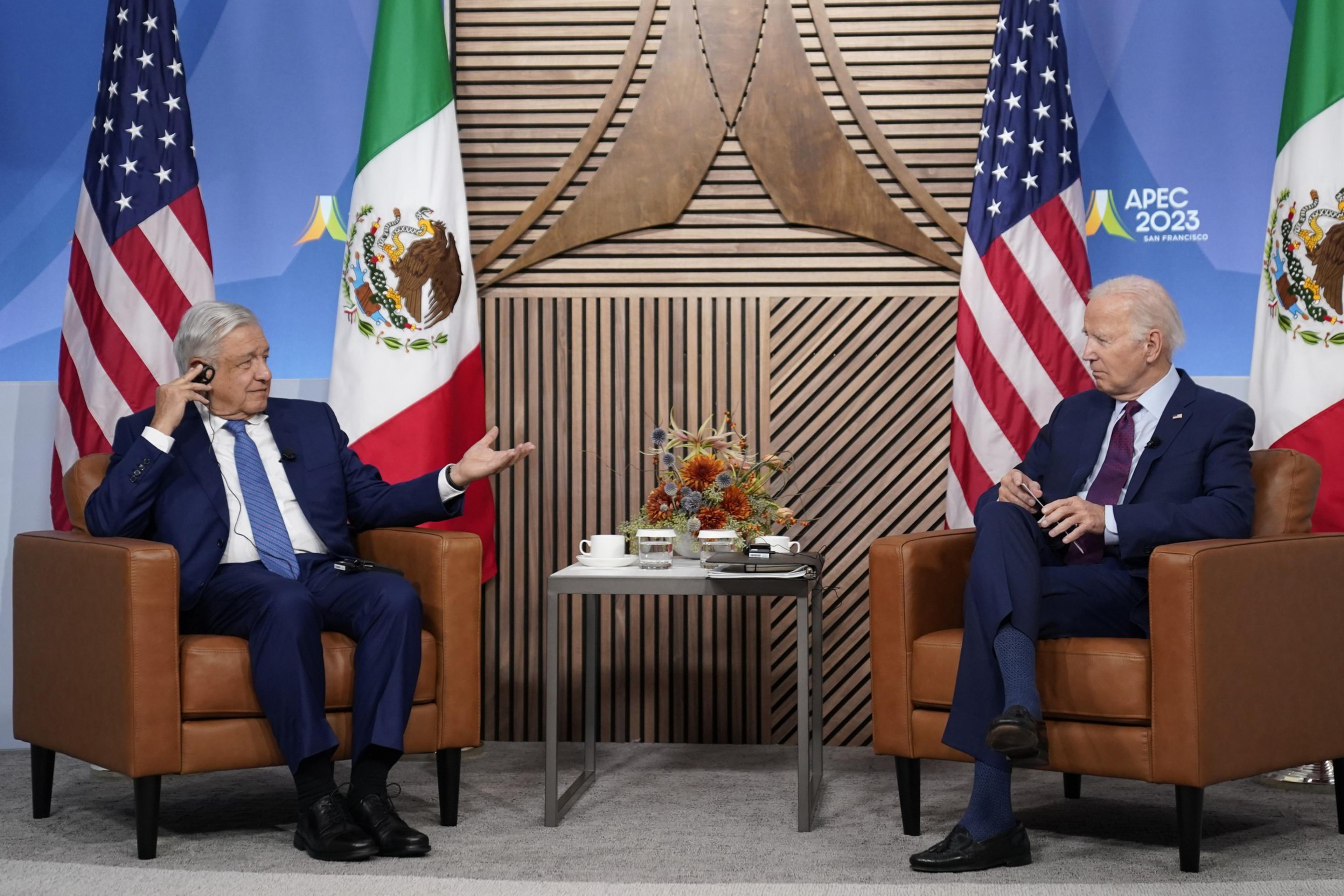 Two men in dark blue suits side in tan chairs with an end table between them. The man on the left sits in front of a Mexican flag. The man on the right, in front of a U.S. flag.