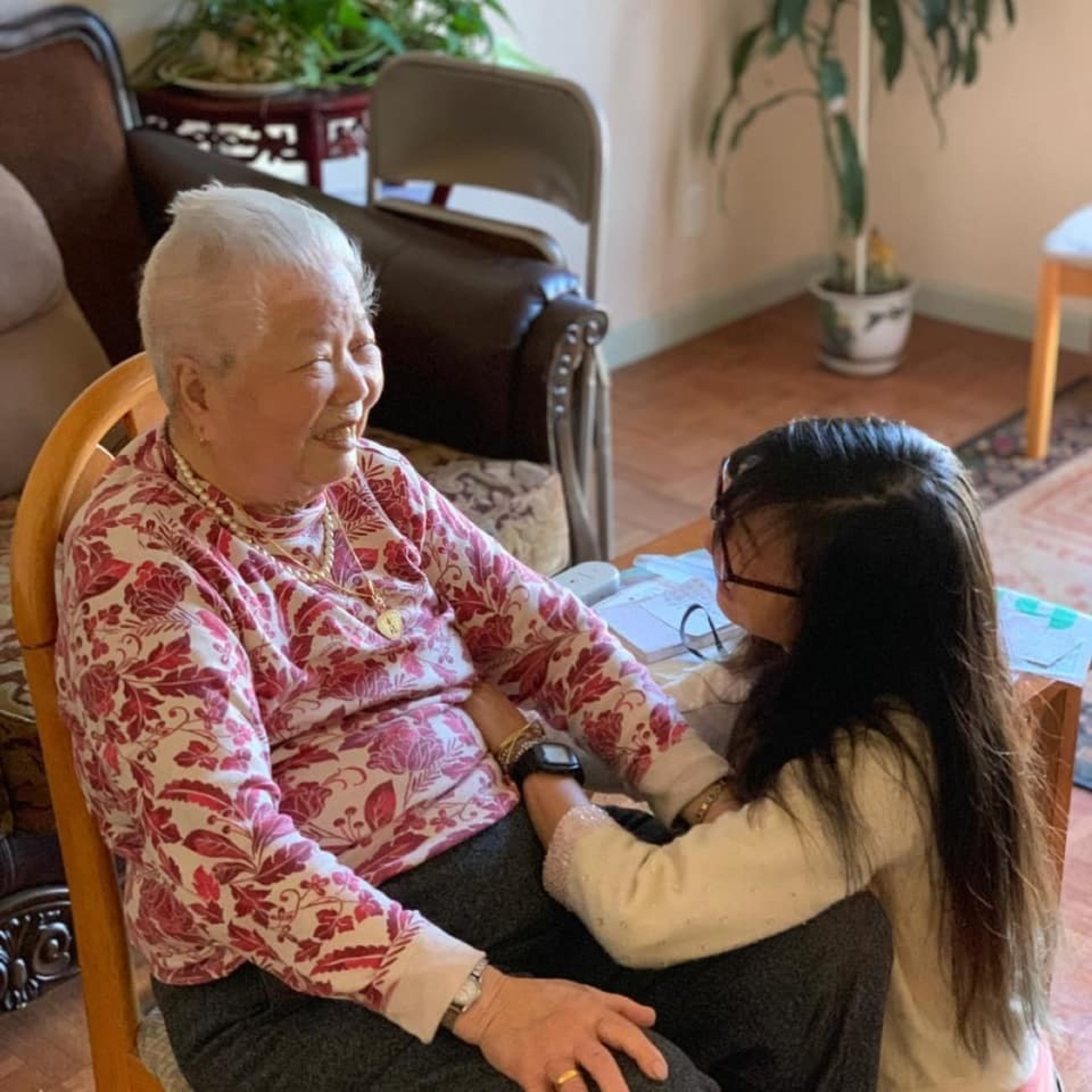 An elderly woman in a pink shirt sits and smiles with her granddaughter at her knee.