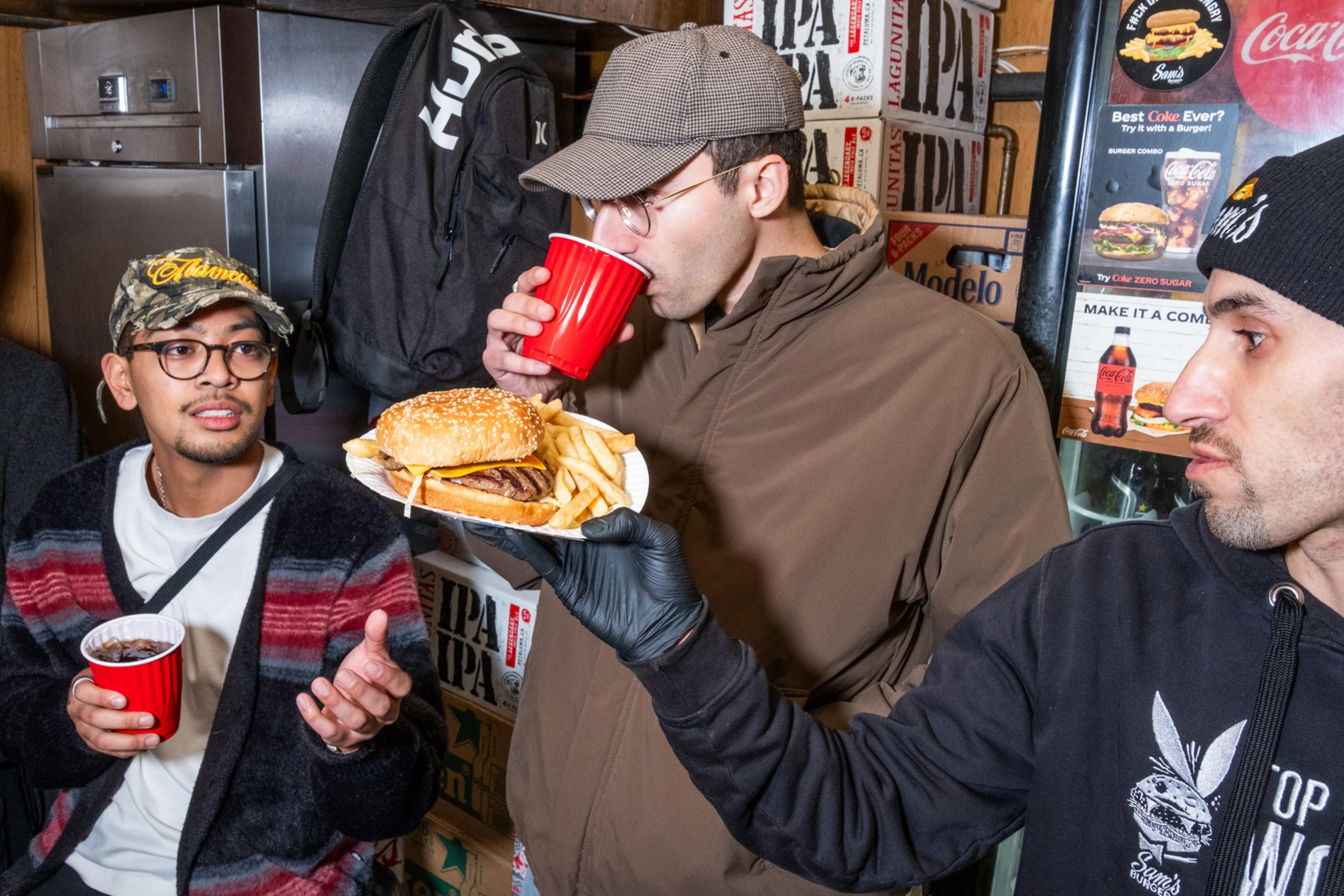 Three people stand together, holding red cups. One person is drinking while another holds a burger and fries on a plate. They are surrounded by beverage boxes.