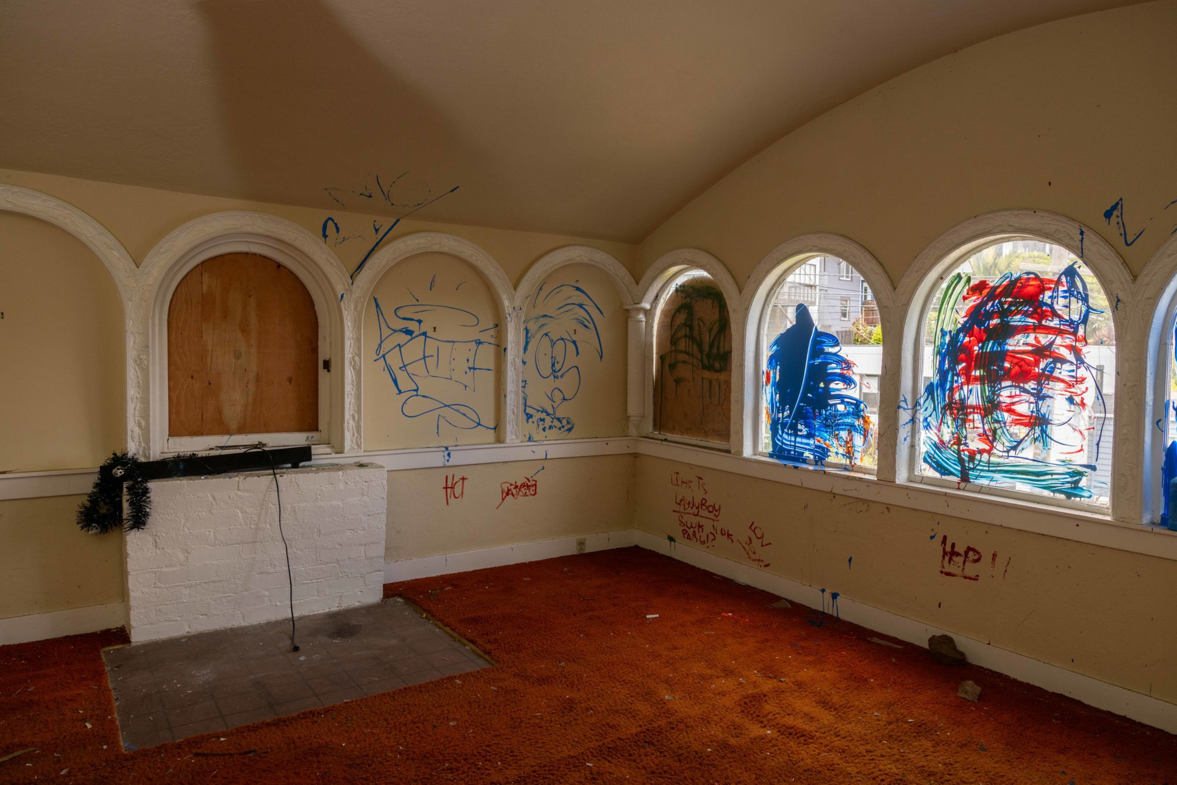 An empty room with red carpet, boarded-up windows, a small white brick fireplace, and walls covered in blue and red graffiti.