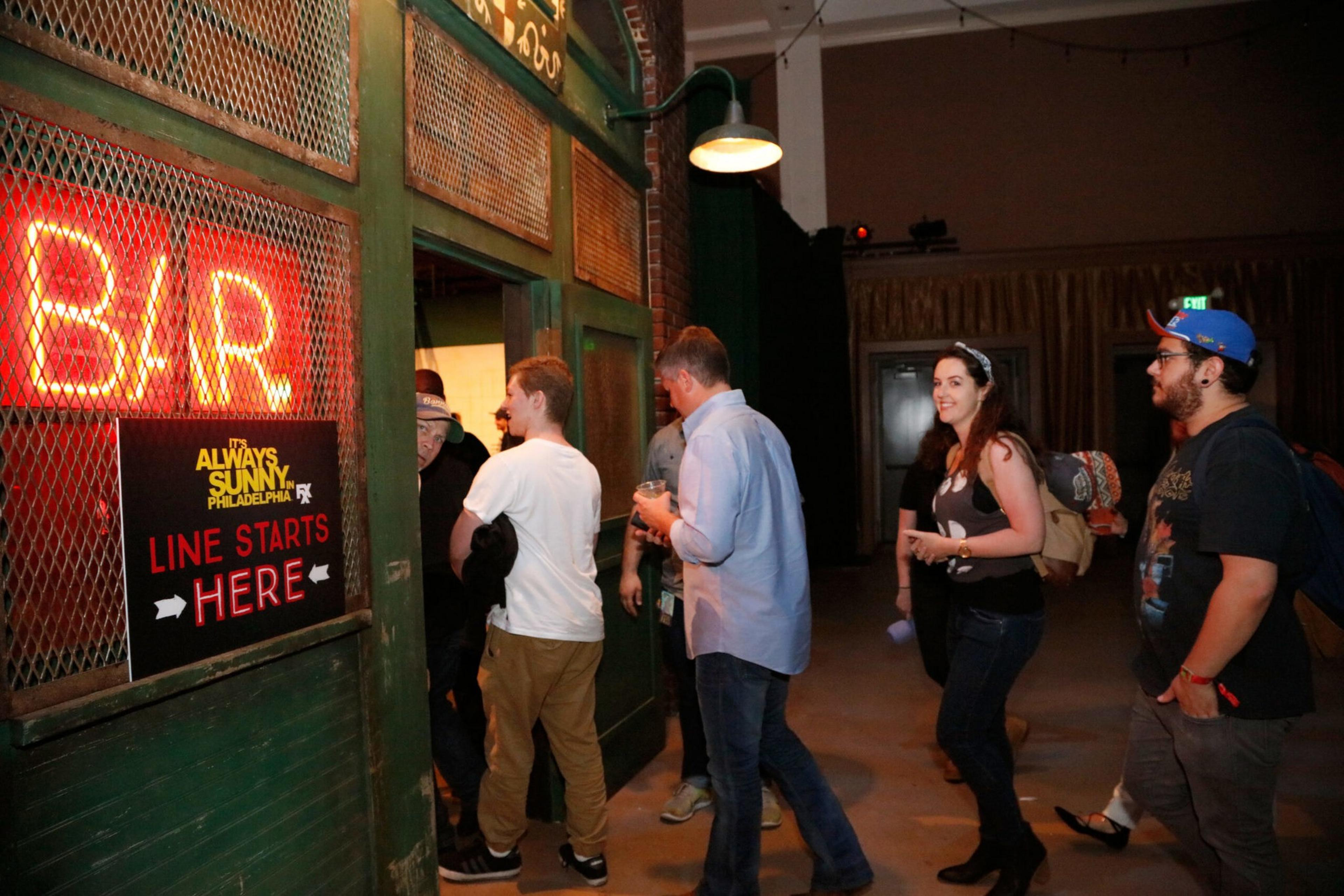 A group of people stand in line inside a dimly lit venue, waiting to enter a bar with a red neon sign and a sign indicating the line starts there.