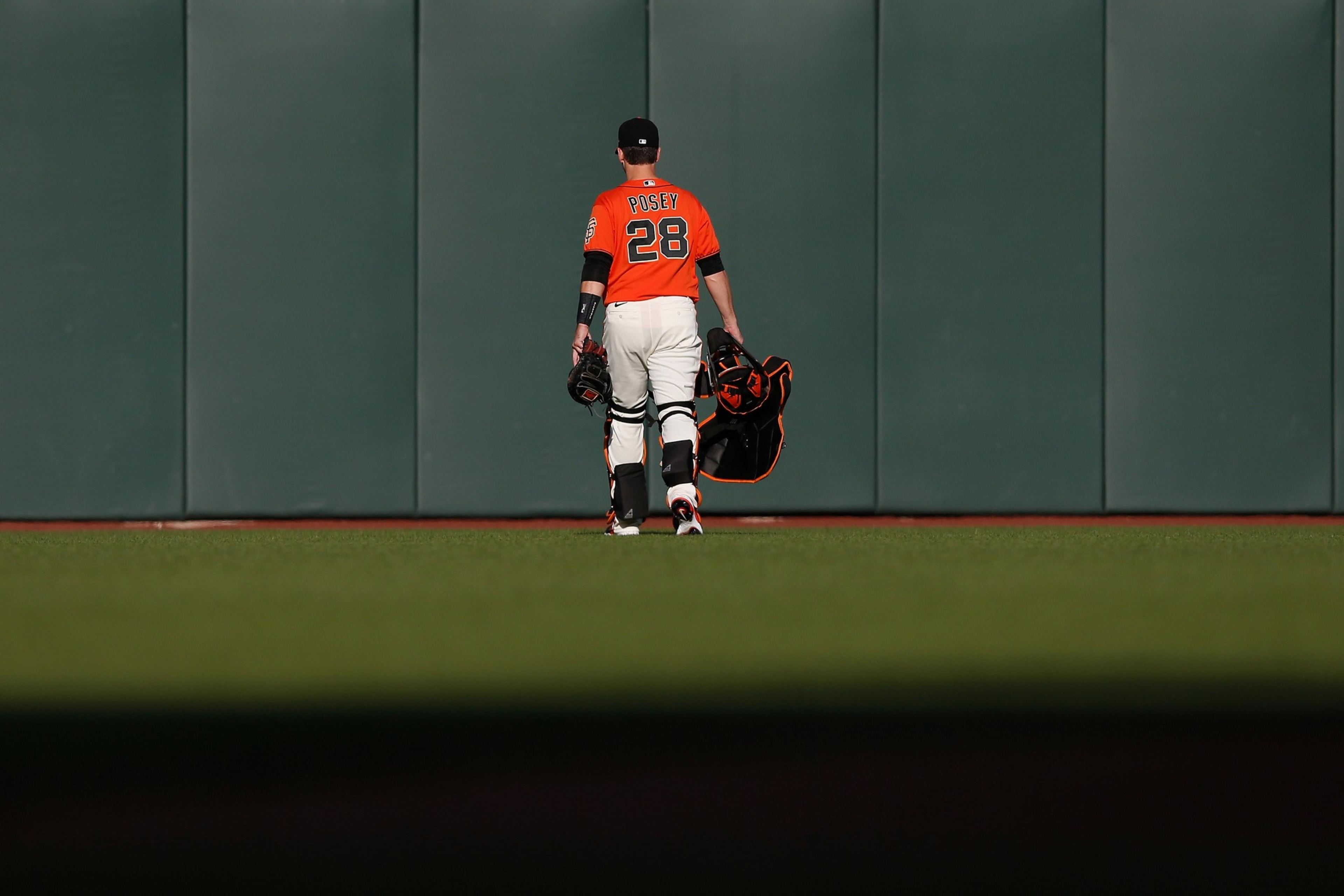 A baseball player in orange and white gear, carrying a helmet and mitt, walks toward a green outfield wall. The player's jersey reads &quot;Posey 28.&quot;