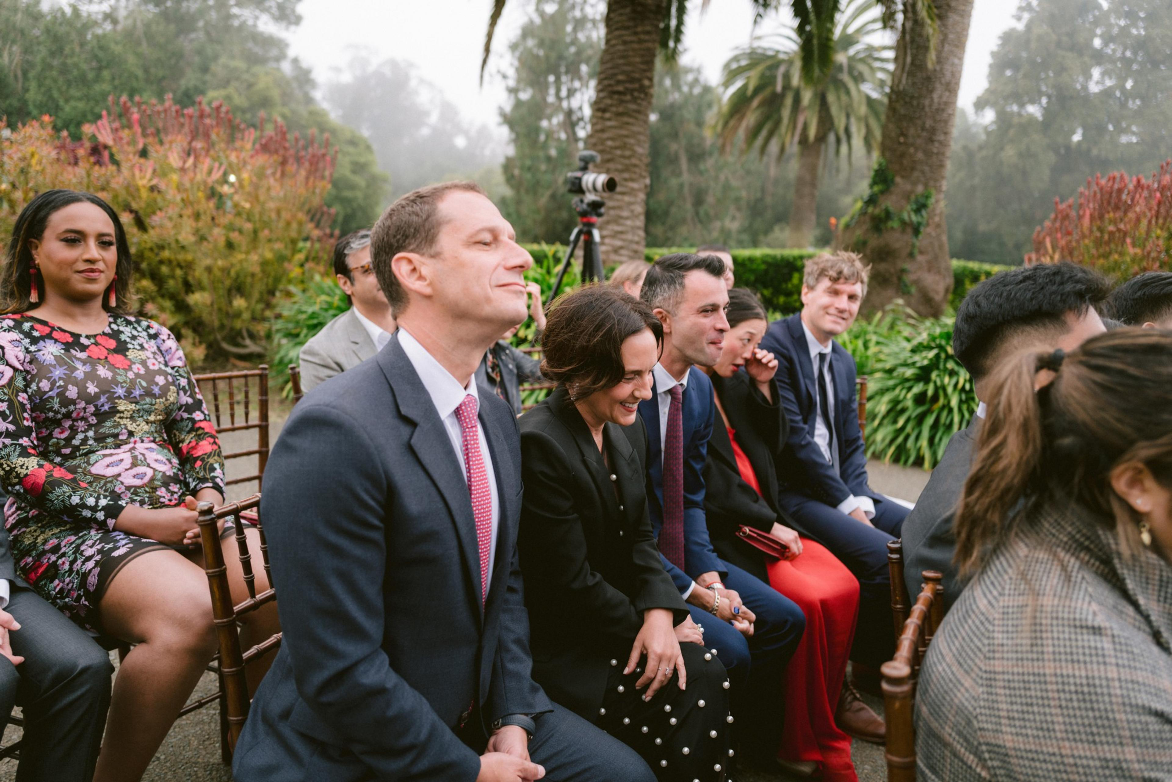A group of people dressed formally are seated outdoors, smiling and chatting, surrounded by greenery and palm trees under a cloudy sky.
