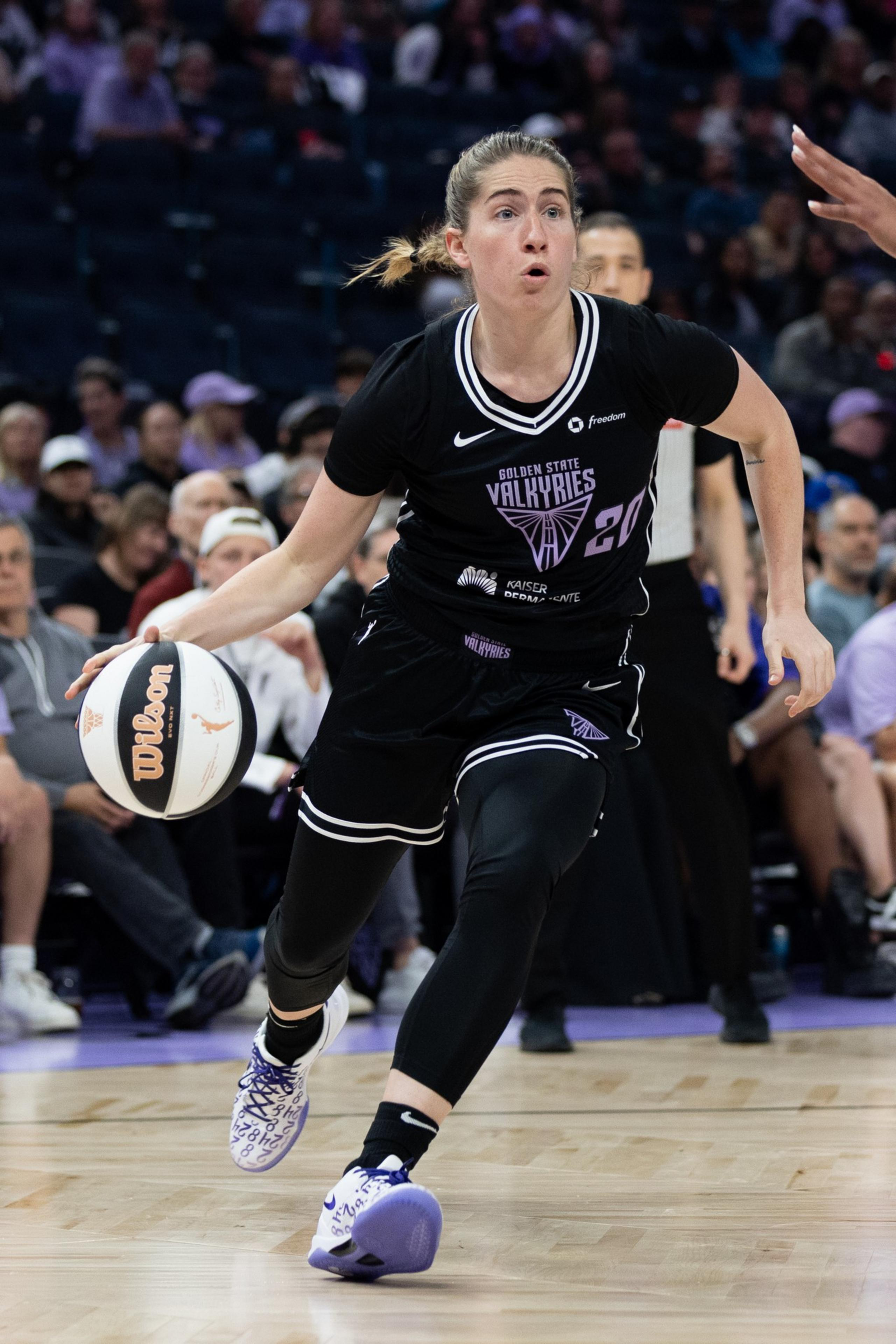 A basketball player in a black &quot;Golden State Valkyries&quot; jersey dribbles the ball on the court, focused and in motion, with an audience in the background.
