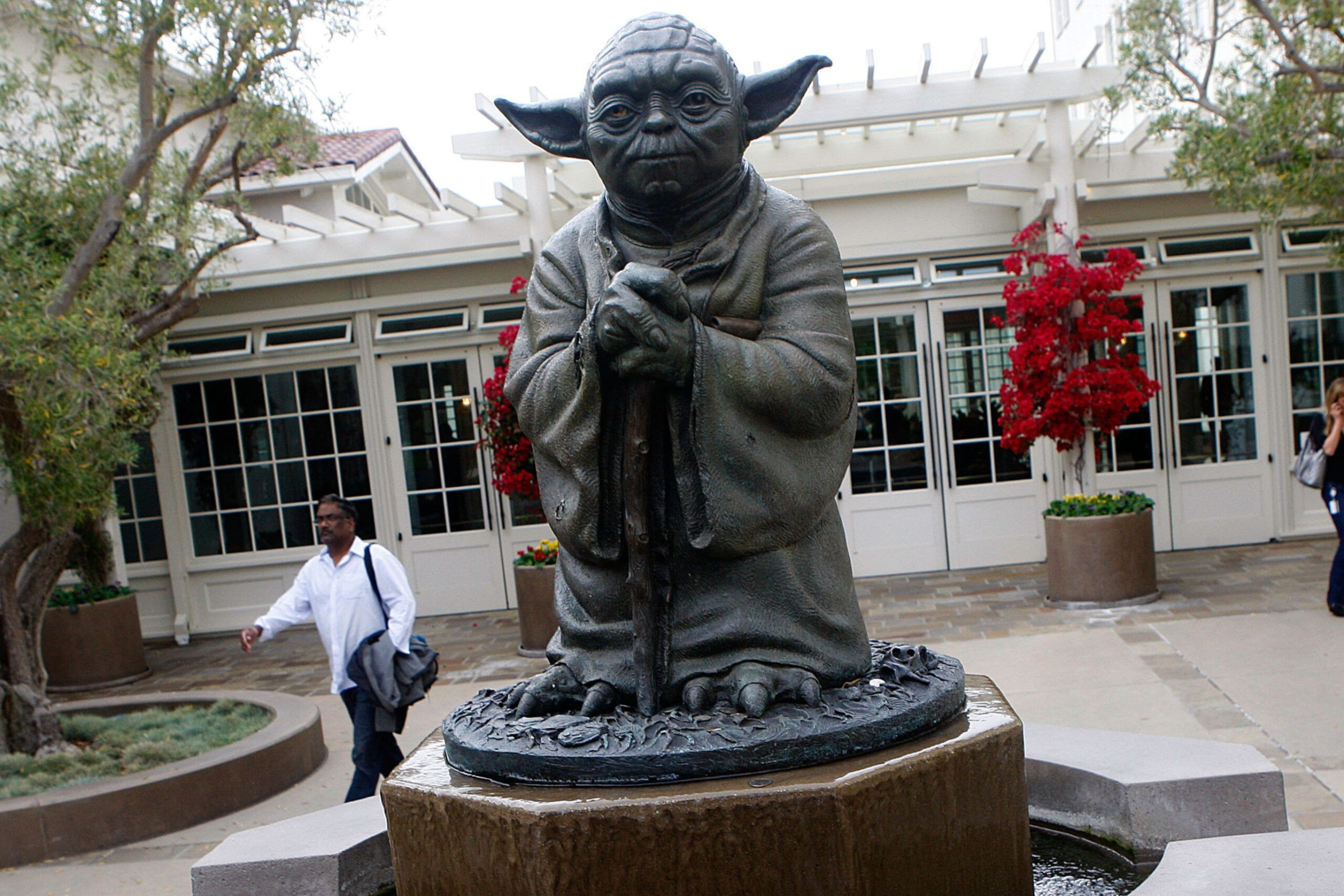 A bronze statue of Yoda holding a walking stick stands on a pedestal in front of a building with large windows and red-leafed trees nearby.