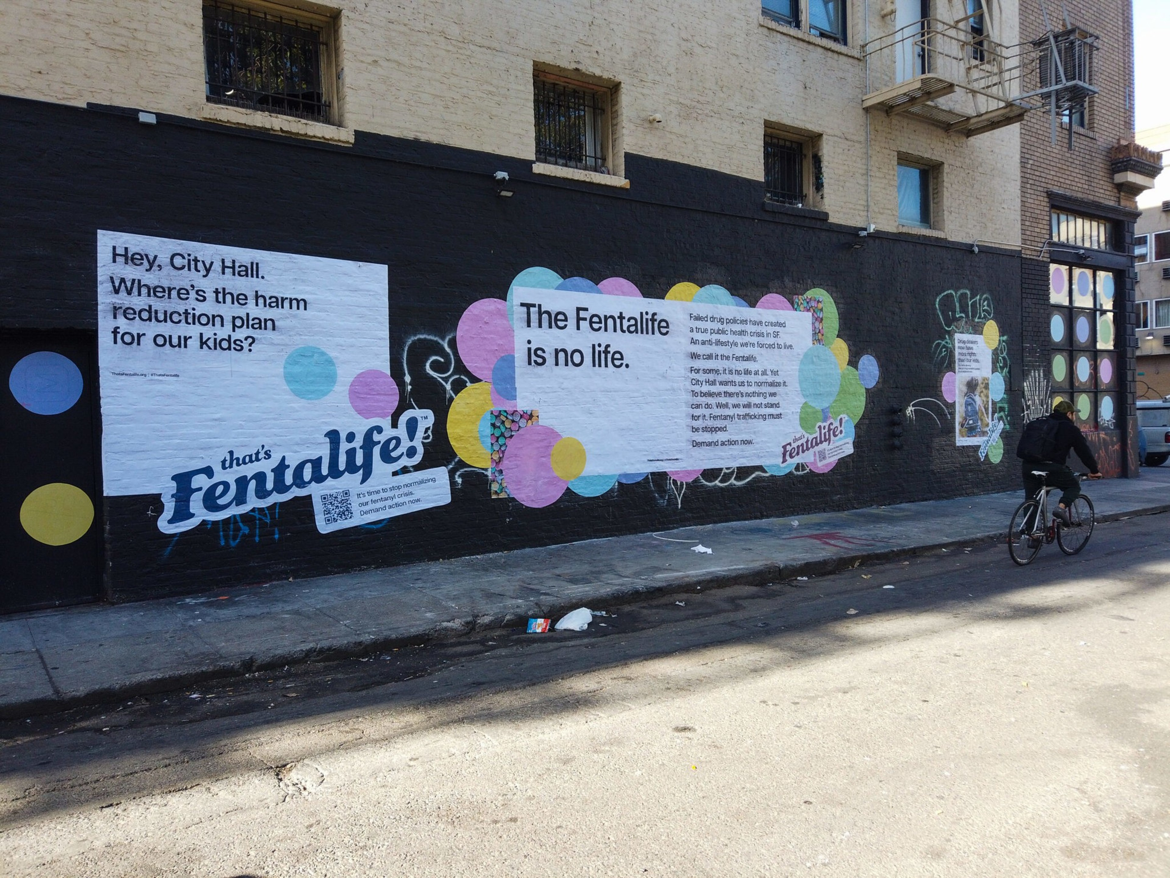 A wall mural displays messages demanding a harm reduction plan for kids, stating "The Fentalife is no life," with colorful circles and a cyclist passing by.