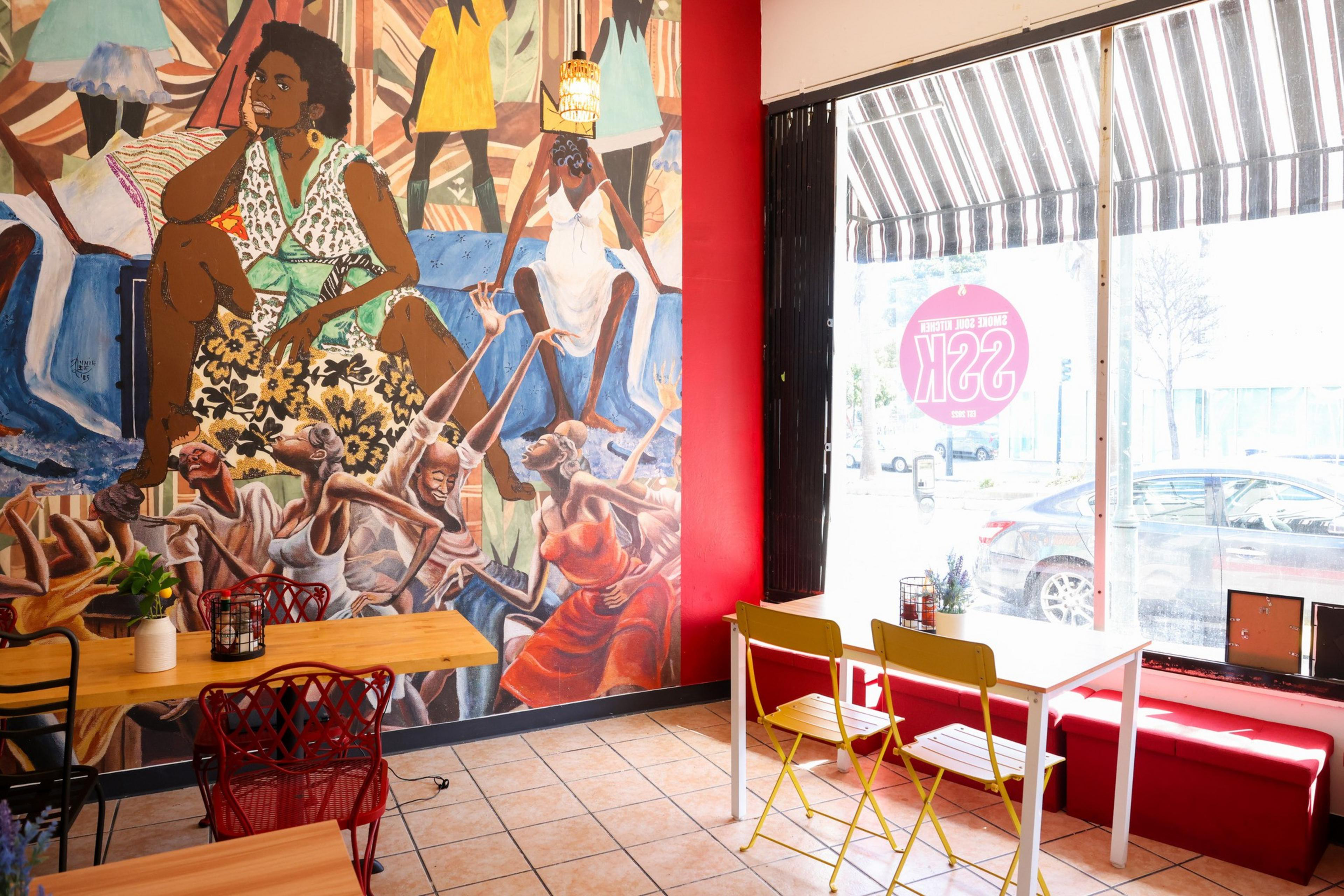 The image shows a vibrant cafe interior with colorful wall art of people, wooden tables, mismatched chairs, and a striped awning outside the window.