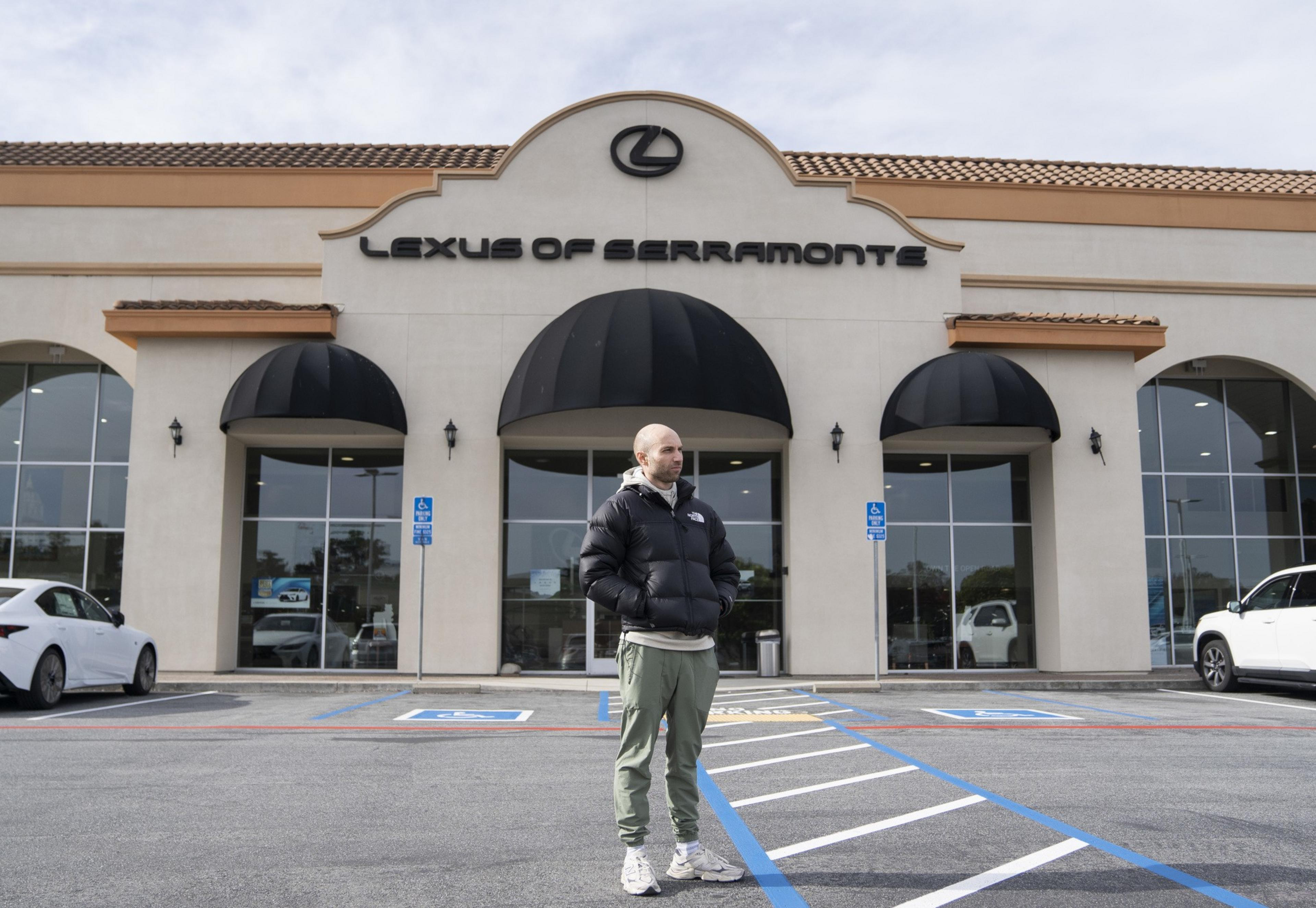 A person in a black puffer jacket and green pants stands outside a &quot;Lexus of Serramonte&quot; dealership with large windows and cars parked nearby.