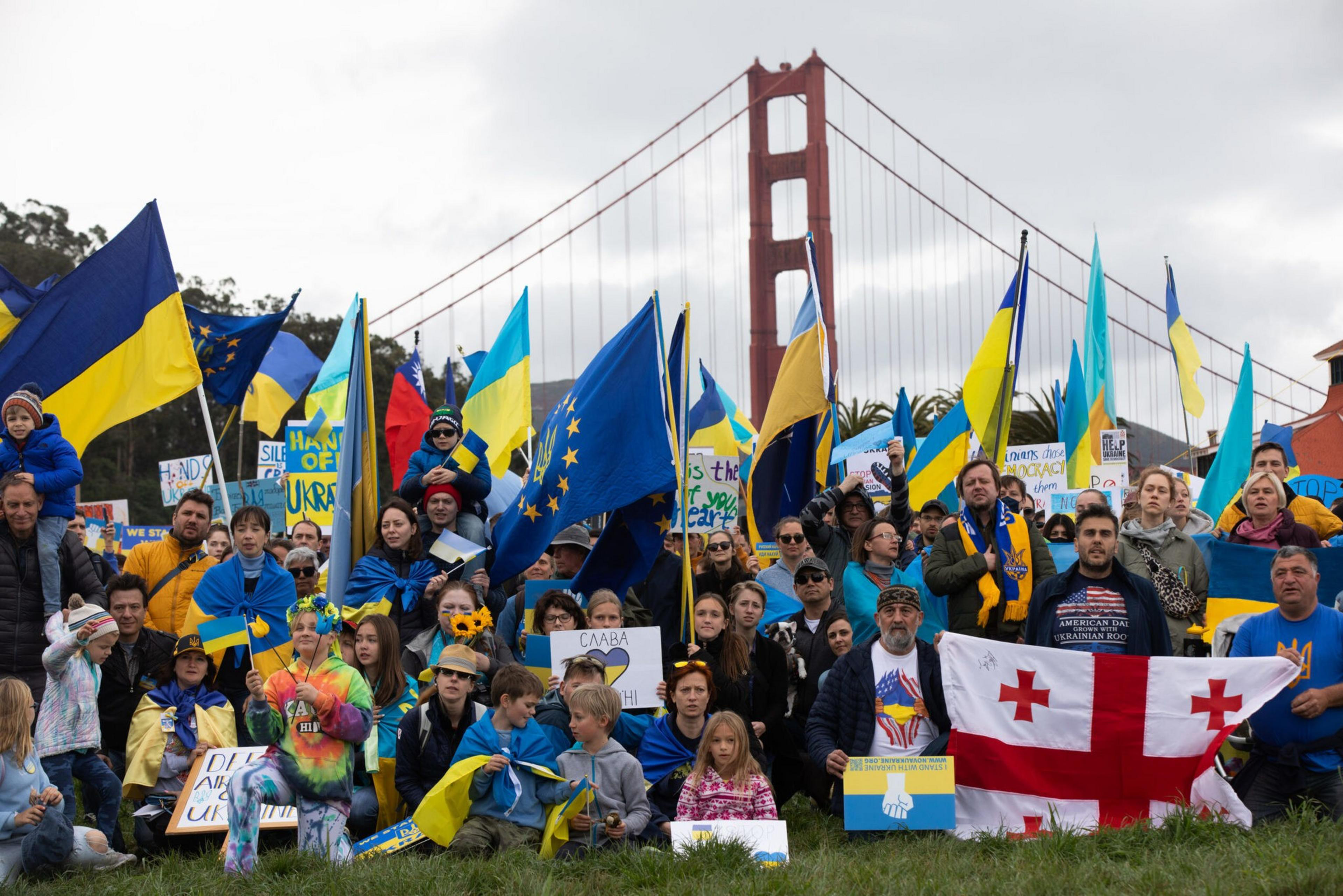 A large group holds Ukrainian and European flags, signs supporting Ukraine, and a Georgian flag, gathered near the Golden Gate Bridge on a cloudy day.
