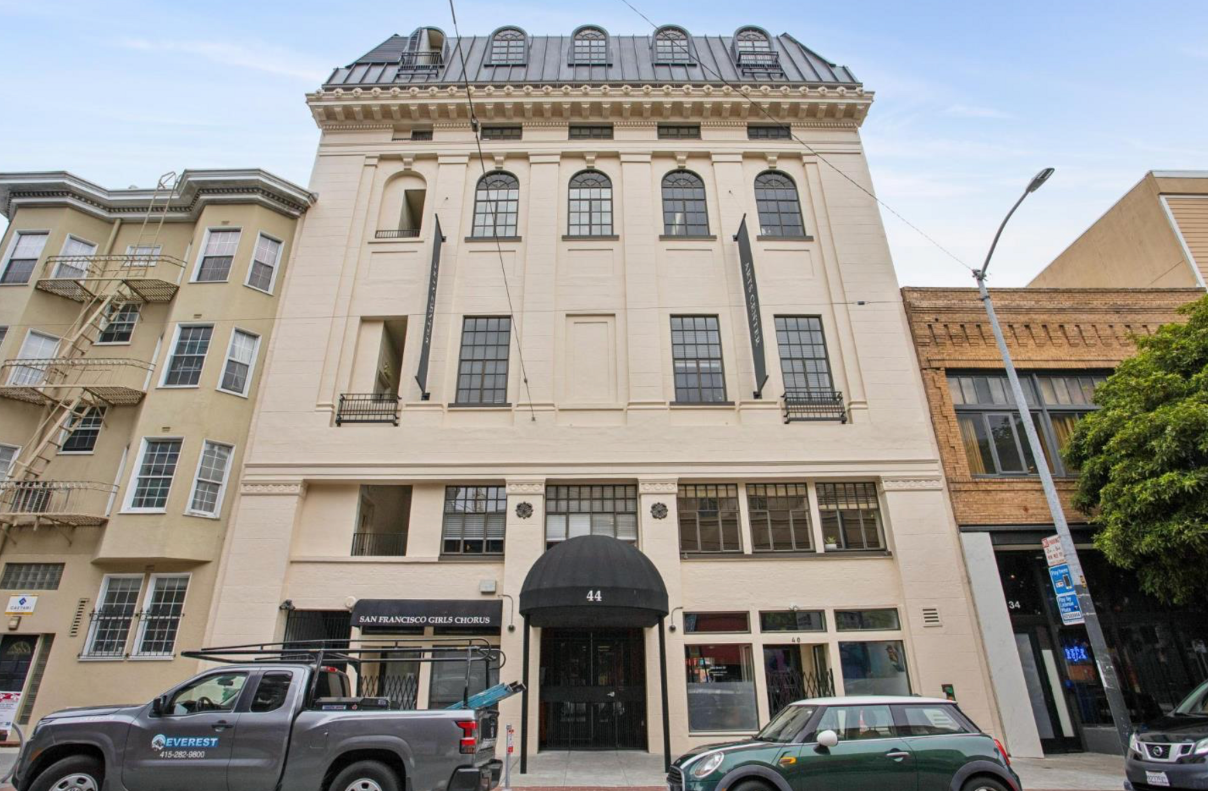 A four-story beige building with arched roof windows and black awnings, a black truck and a green car parked in front on the street.