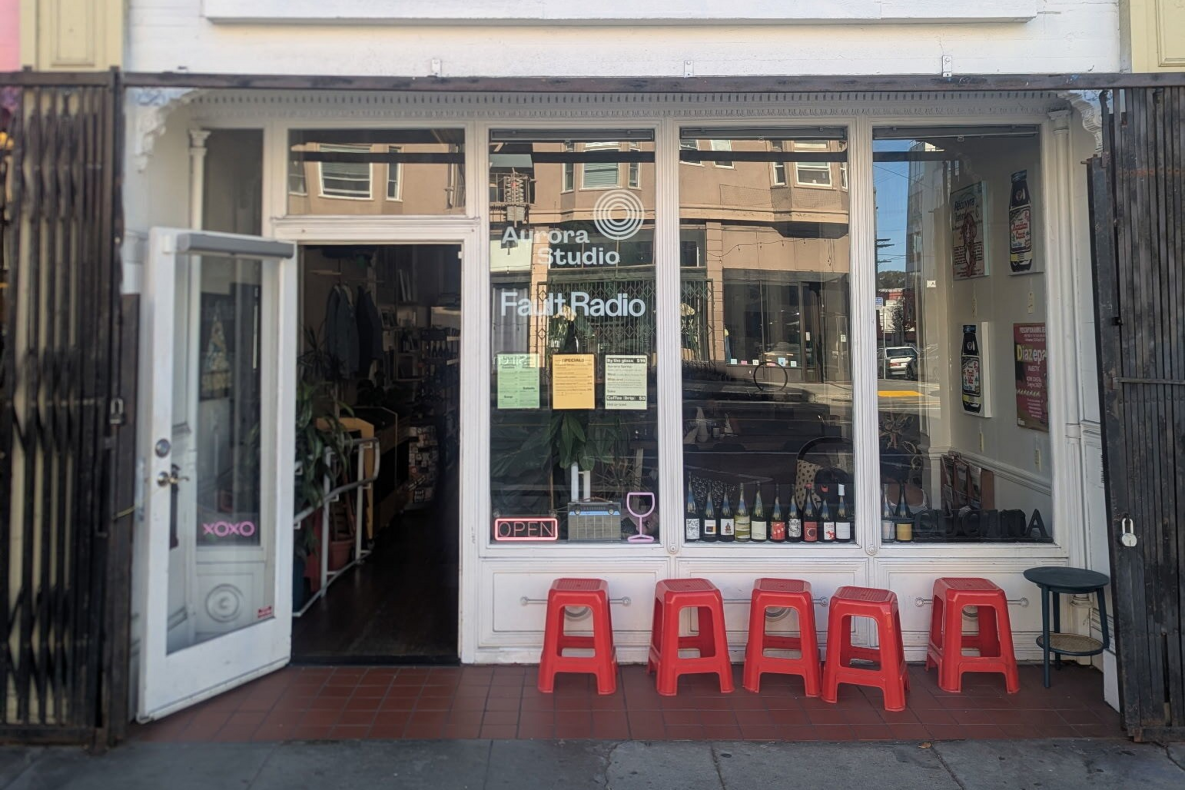 A storefront with glass windows and an open door displays "Aurora Studio" and "Fault Radio," with five red stools outside and various bottles lined up in the window.