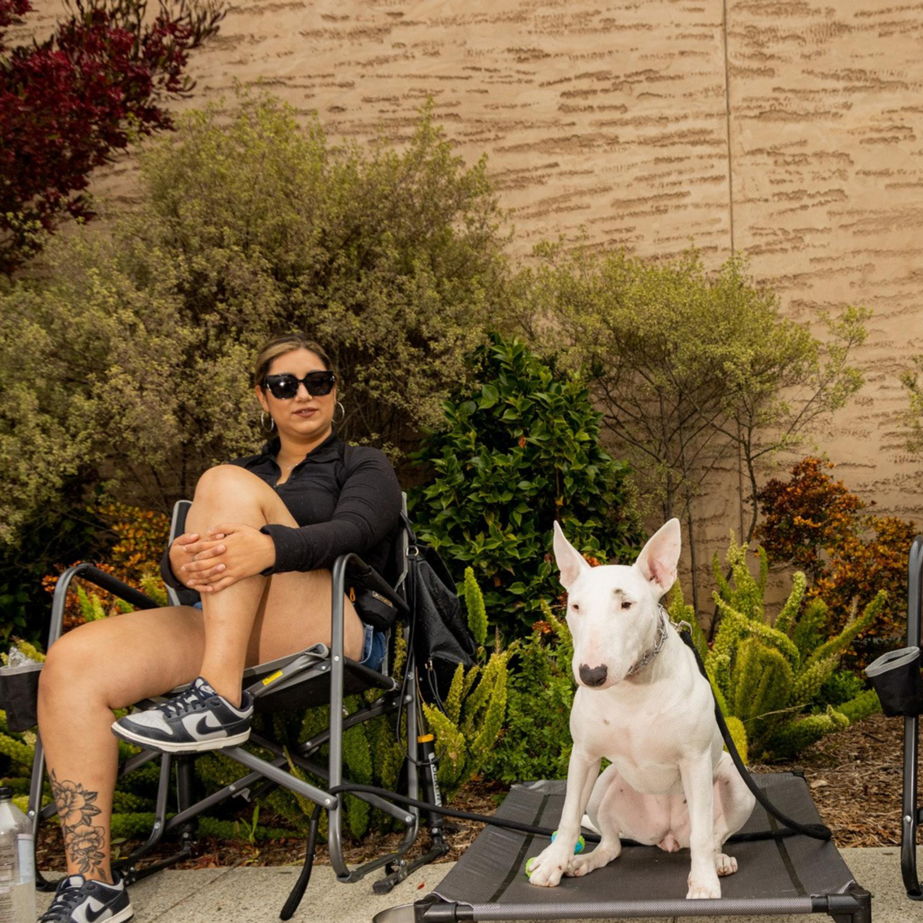 Two adults and a child sit near bushes, with a white dog on a raised bed between the adults. The adults are relaxed on chairs, and the child stands nearby.