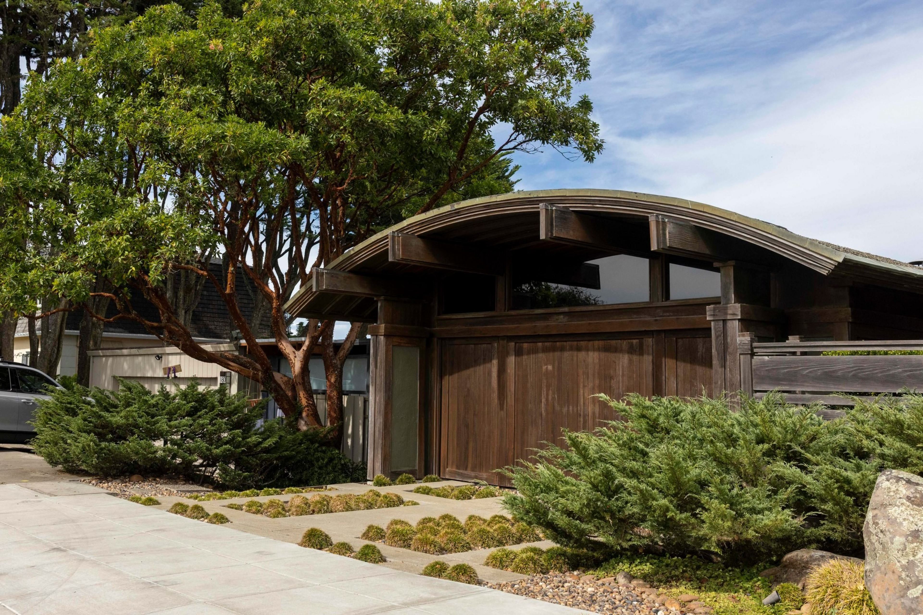 A modern wooden garage with a curved roof sits beside a driveway, surrounded by green shrubs, small mossy patches, and a leafy tree.