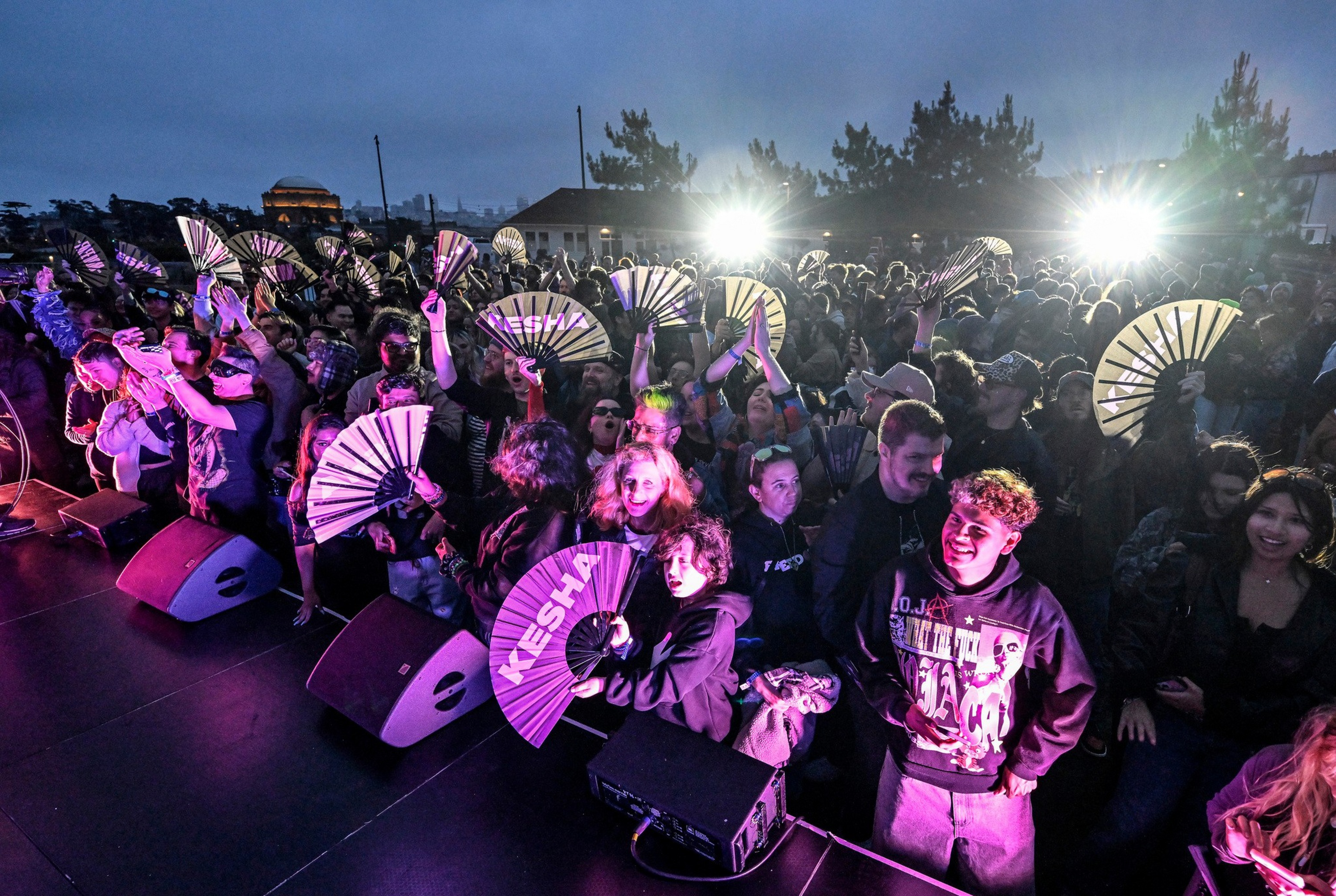 A crowd at a nighttime event holds up fans, with one fan reading “KESHA,” while bright lights illuminate the group from behind.