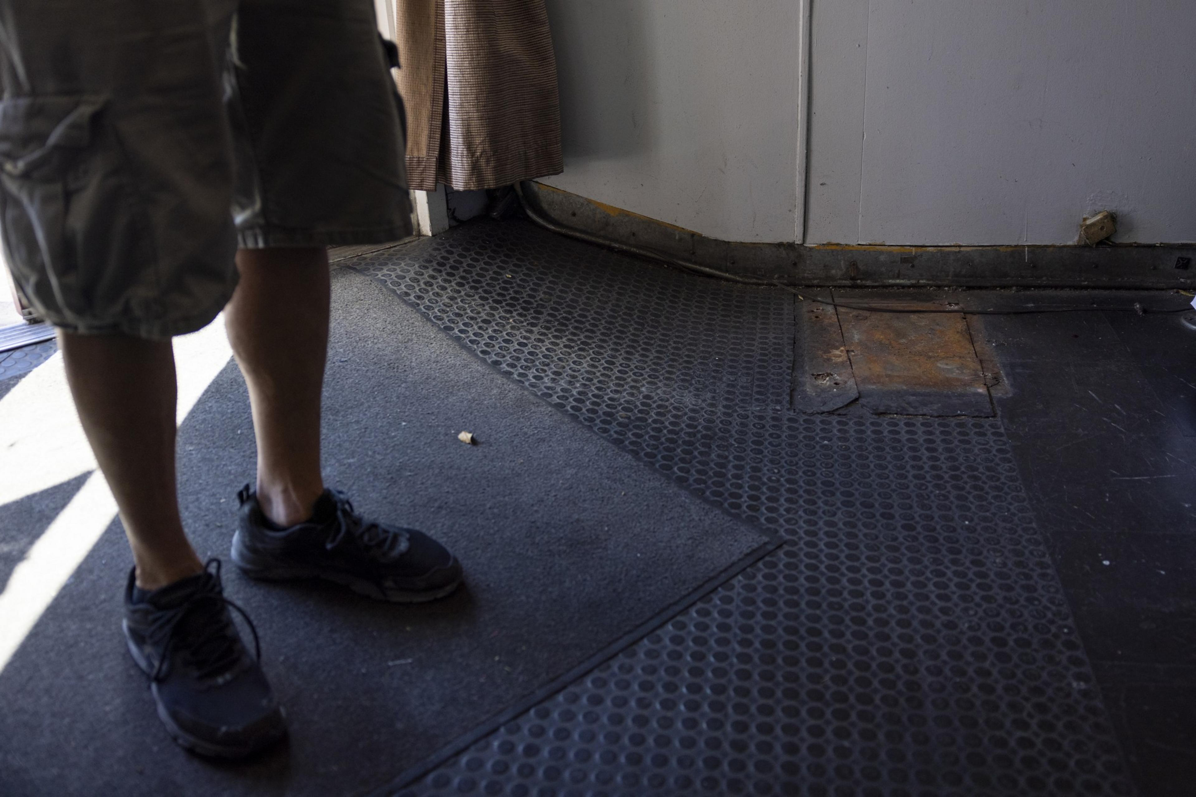 The image shows the lower half of a person wearing cargo shorts and black sneakers, standing on a checkerboard-patterned rubber mat in front of a door with a rusty metal threshold.