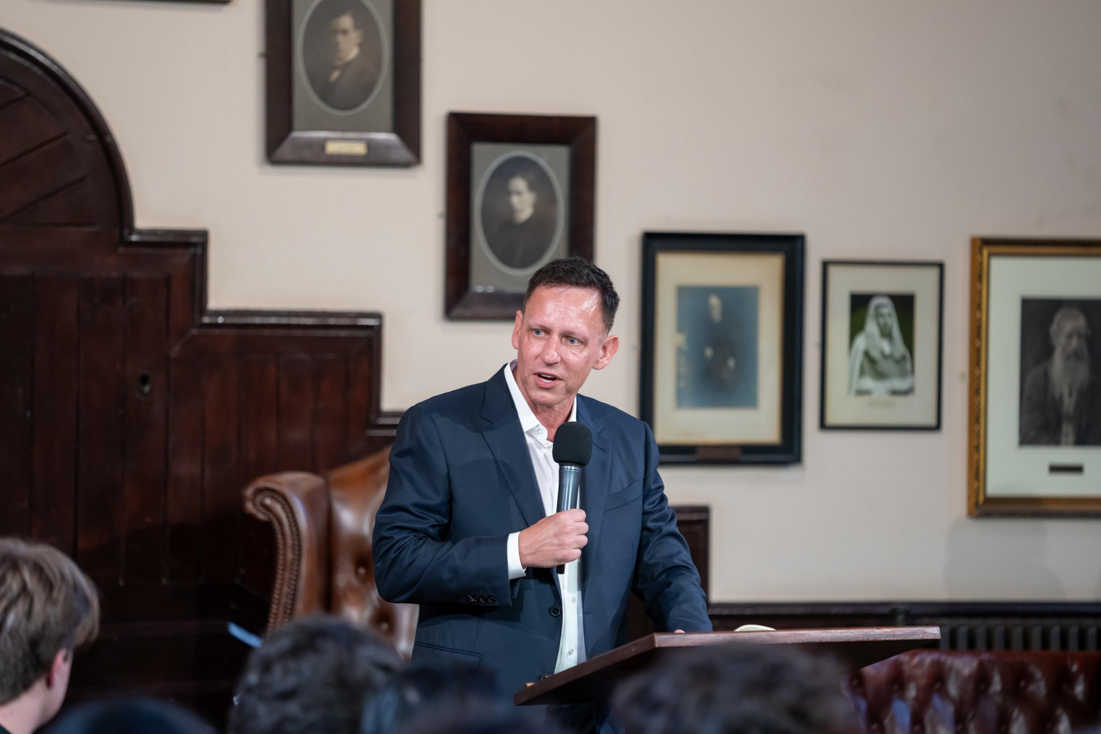 A man in a dark suit holds a microphone and speaks at a podium in a room decorated with framed black-and-white portraits on the wall.