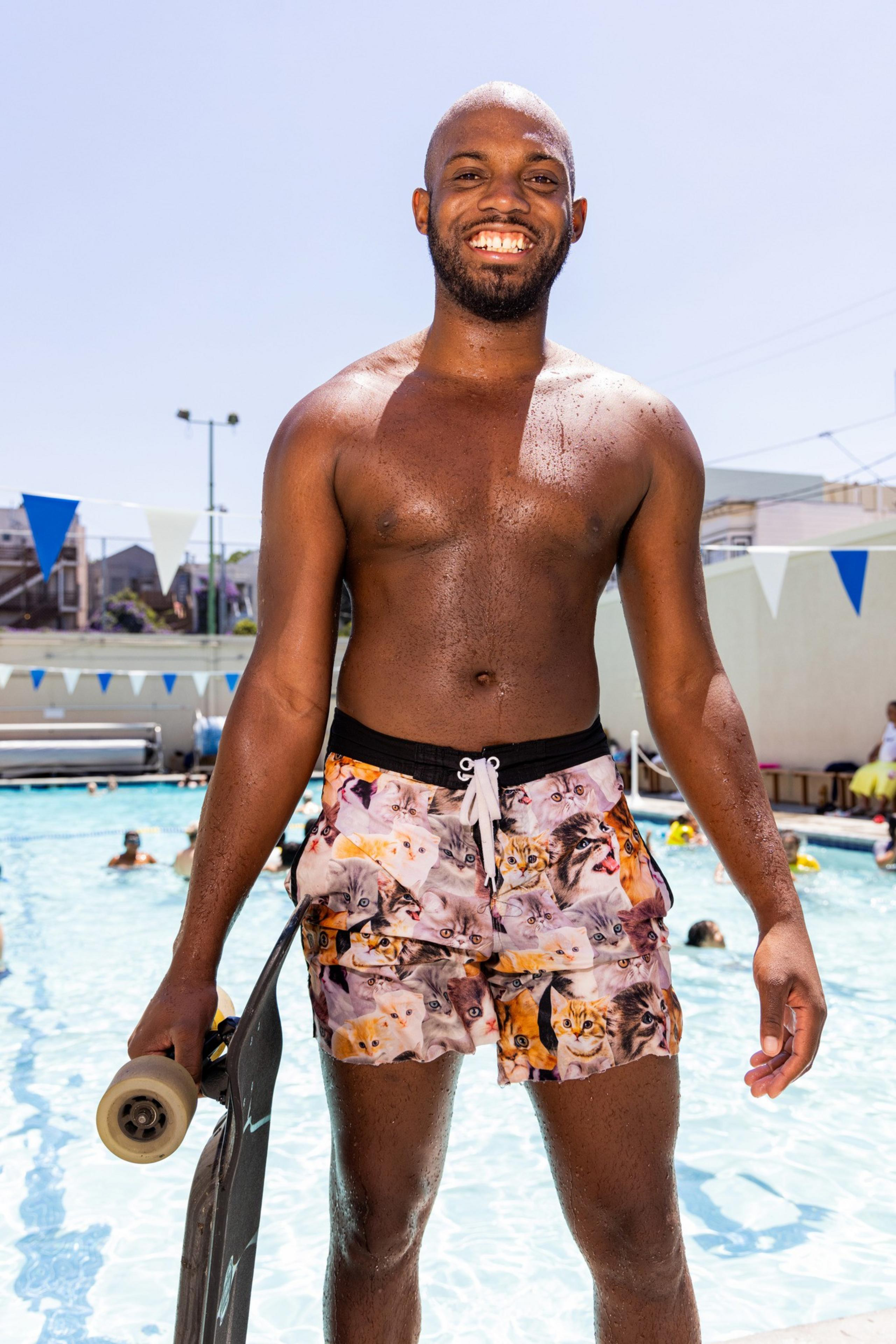 A smiling man stands beside a pool, holding a skateboard. He is shirtless, wearing colorful swim shorts with cat faces printed on them. People are swimming in the background.