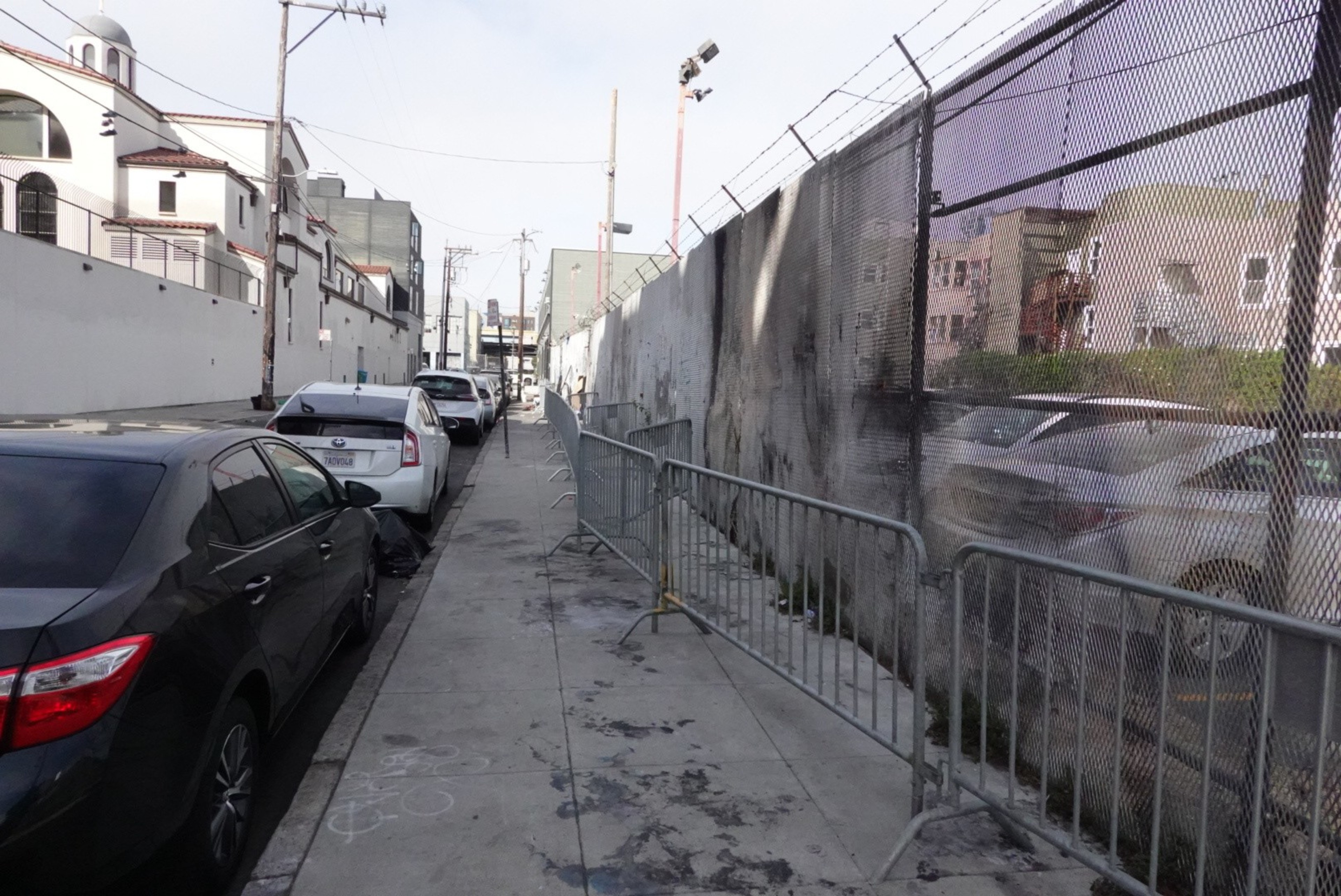The image shows a narrow sidewalk with metal barriers and a fence on one side, parked cars on the street, and a row of buildings lining the other side.